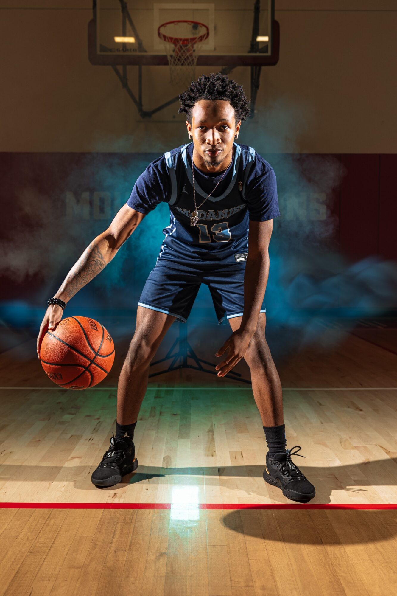 Young man dribbling basketball on court with colorful smoke background, focused stance, wearing sportswear and sneakers.