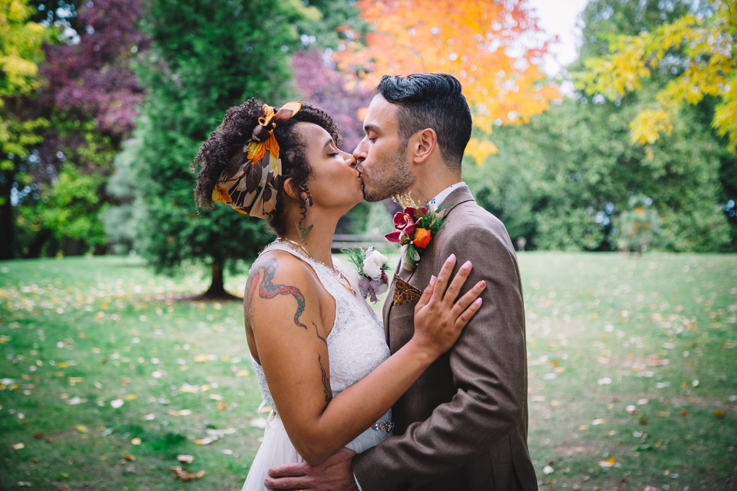 Couple kissing outdoors in a park with colorful autumn trees in the background.