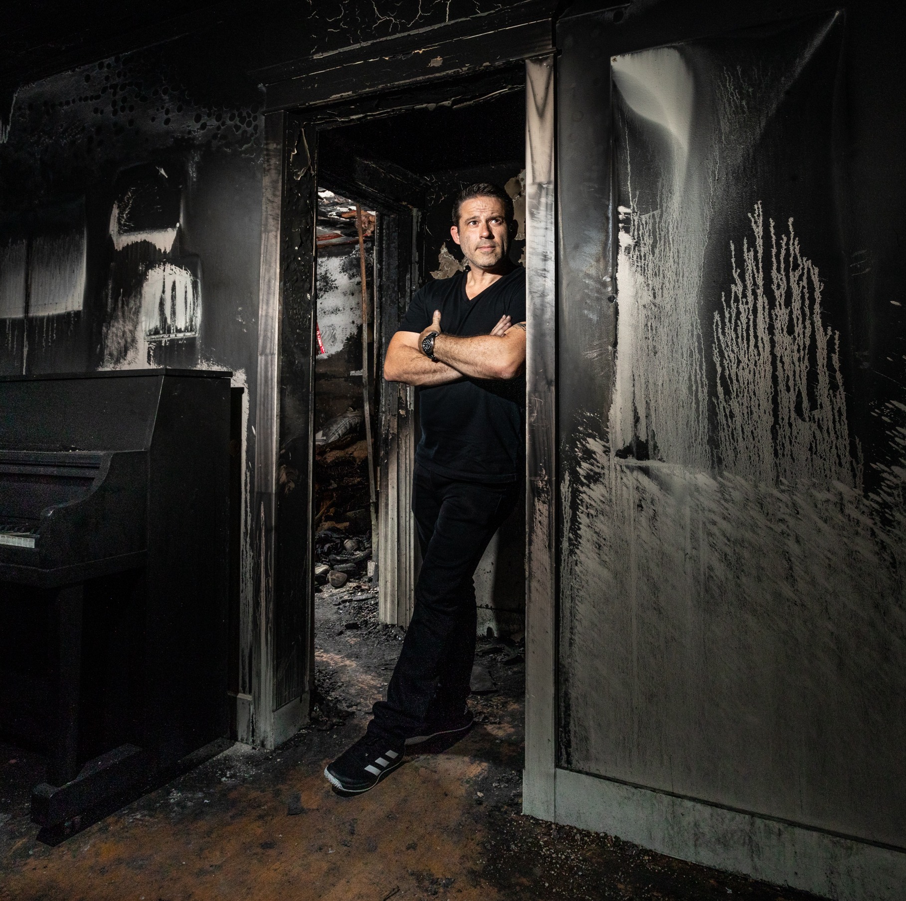 Man standing in doorway of burnt room with blackened walls and soot, arms crossed, looking at camera.