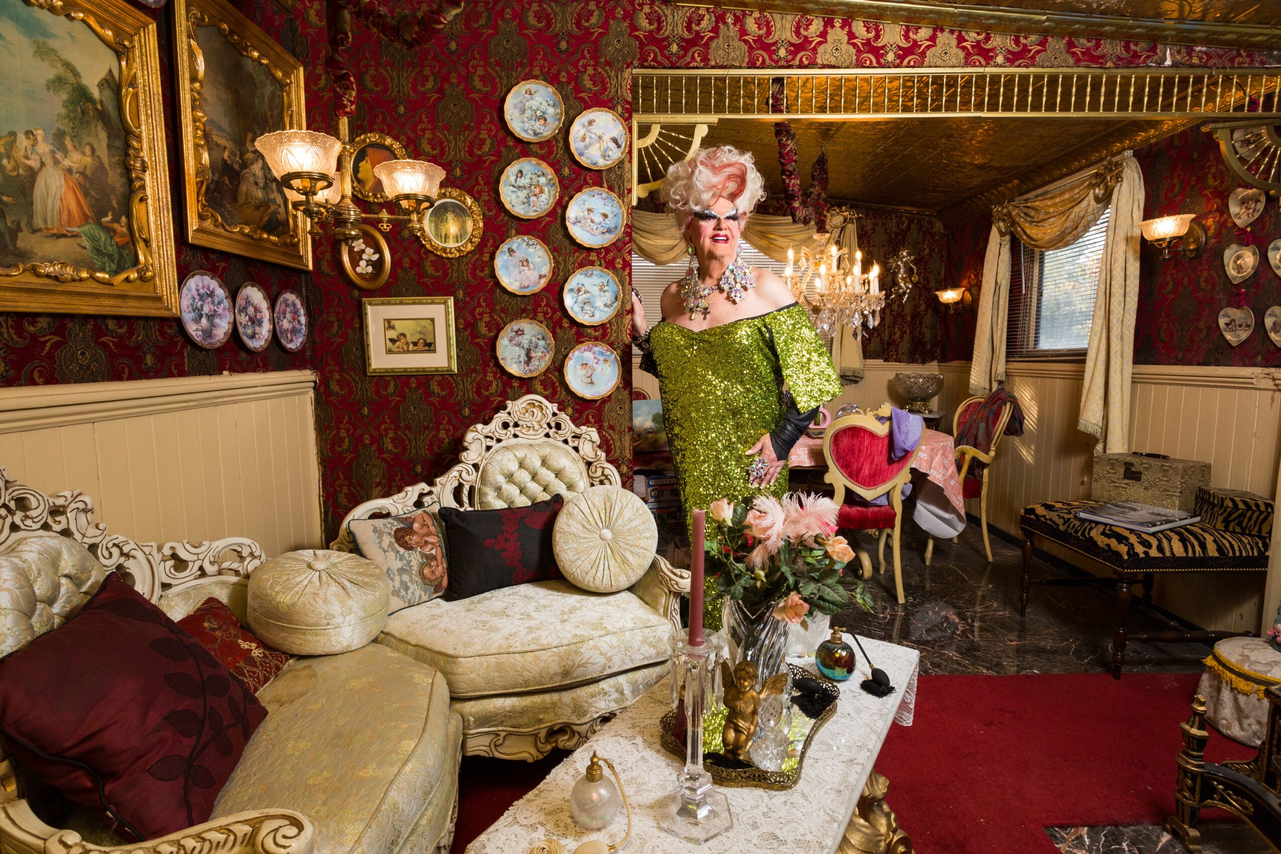 Woman in green dress with floral headpiece standing in a vintage decorated room with sofas, plates, and paintings