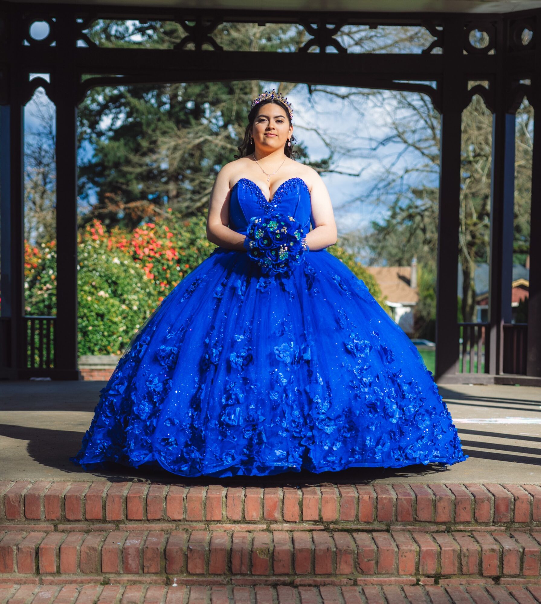 Young woman in a blue ball gown holding a bouquet, standing on a brick platform under a gazebo, outdoors.