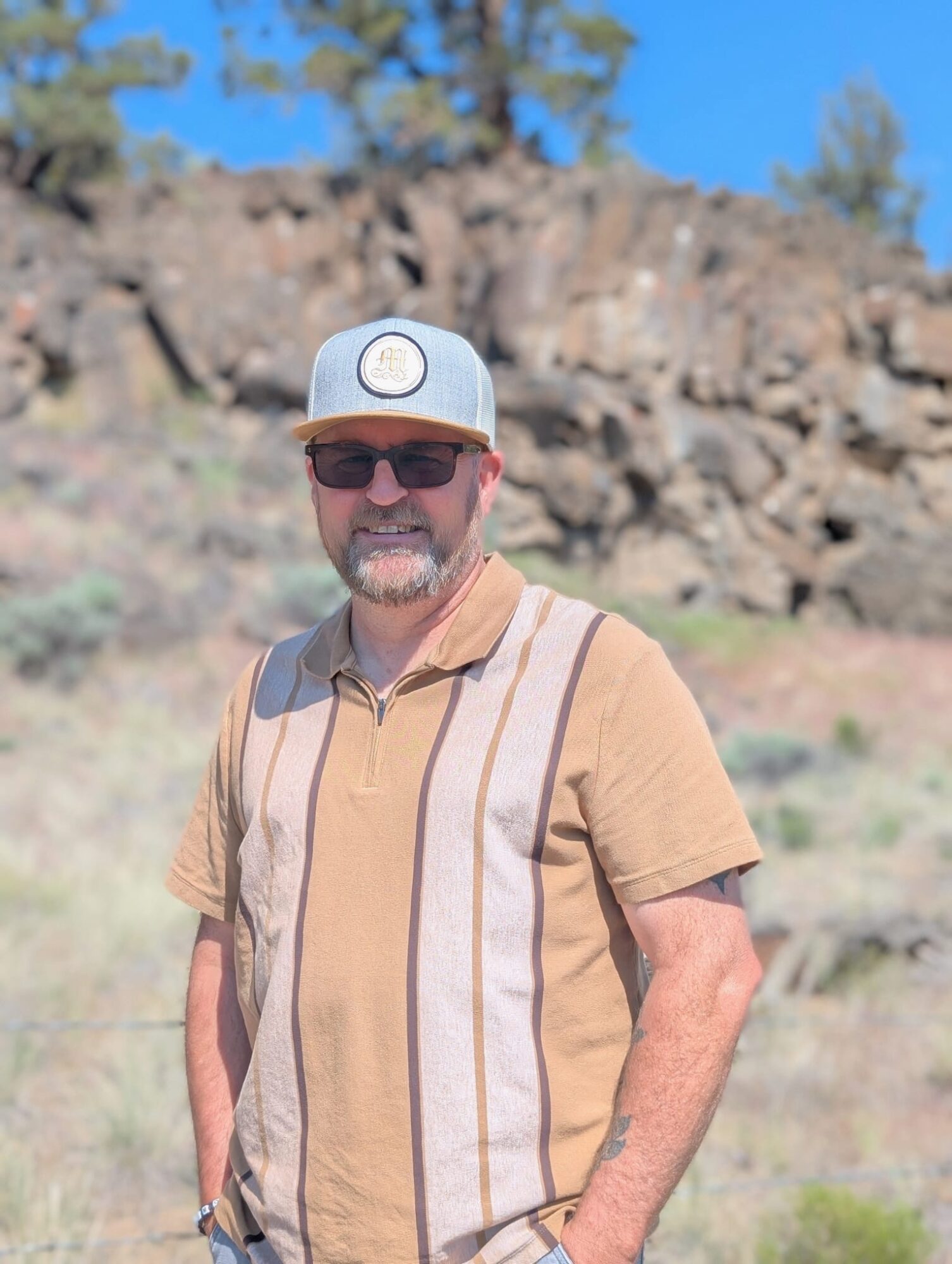 Man wearing sunglasses, a cap, and a tan striped shirt outdoors with rocks and trees in background.