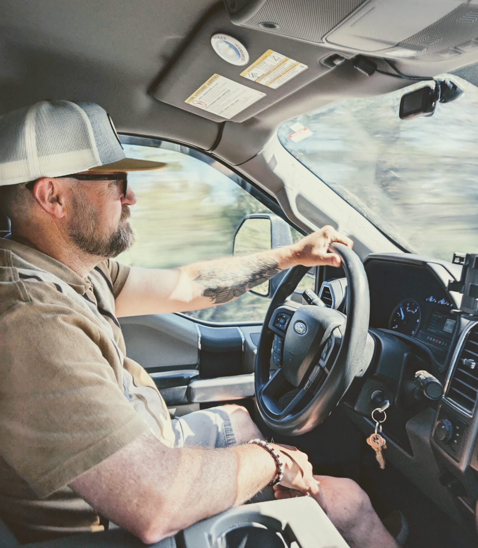 Man with a beard and sunglasses driving a vehicle, wearing a cap and casual clothing, with a view of the road outside.