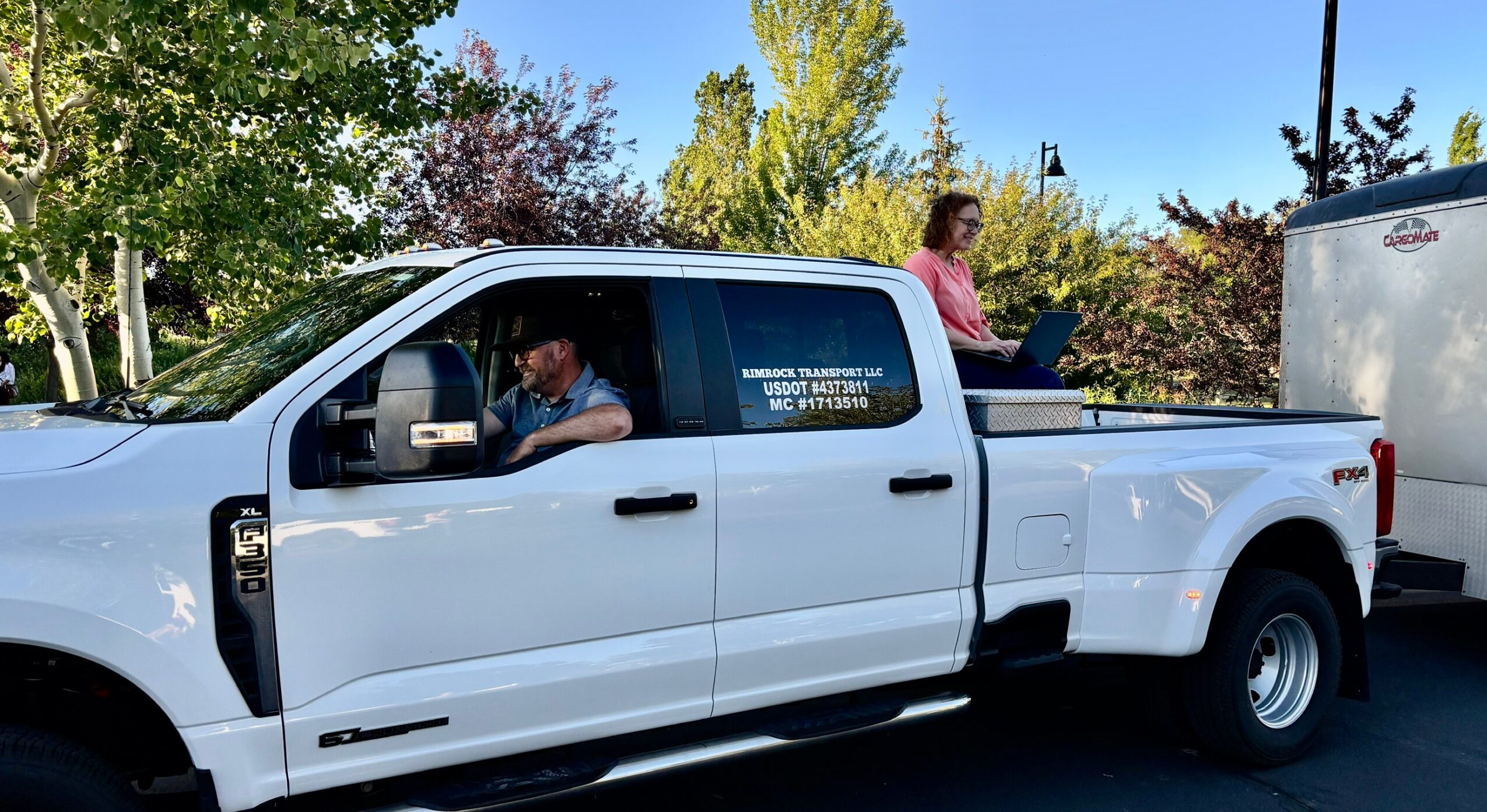 White pickup truck with two people inside, one in the driver's seat and one in the back, parked outdoors with trees and a clear sky.