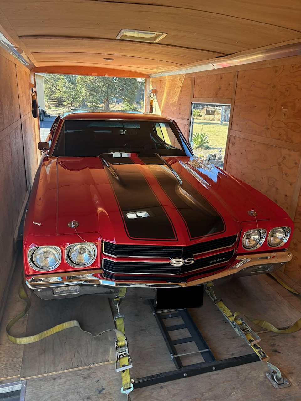 Red classic Chevrolet SS car with black racing stripe inside a wooden garage, viewed from front, with sunlight outside.
