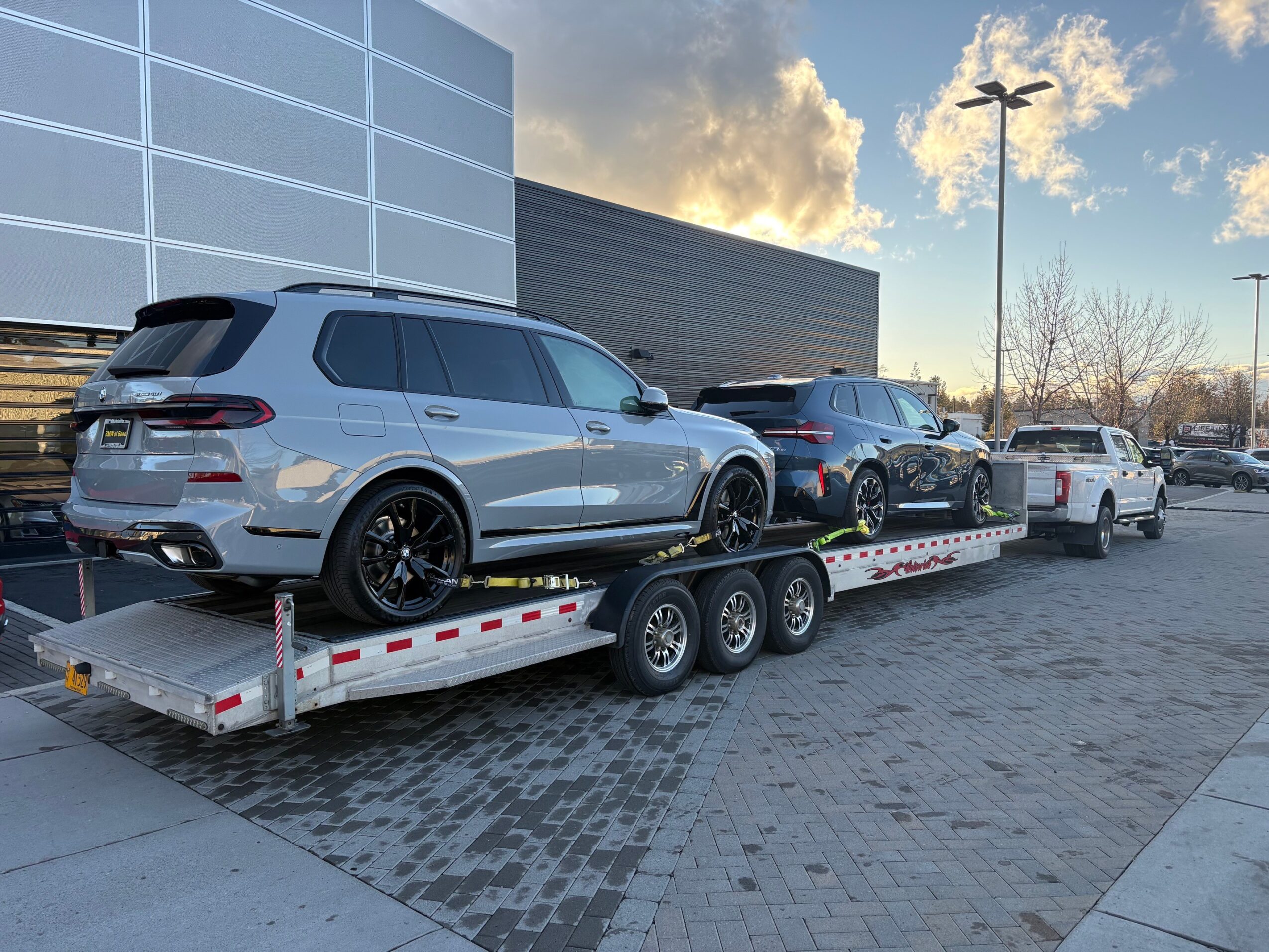 Silver SUV on a flatbed trailer in parking lot with other cars and modern building in background.