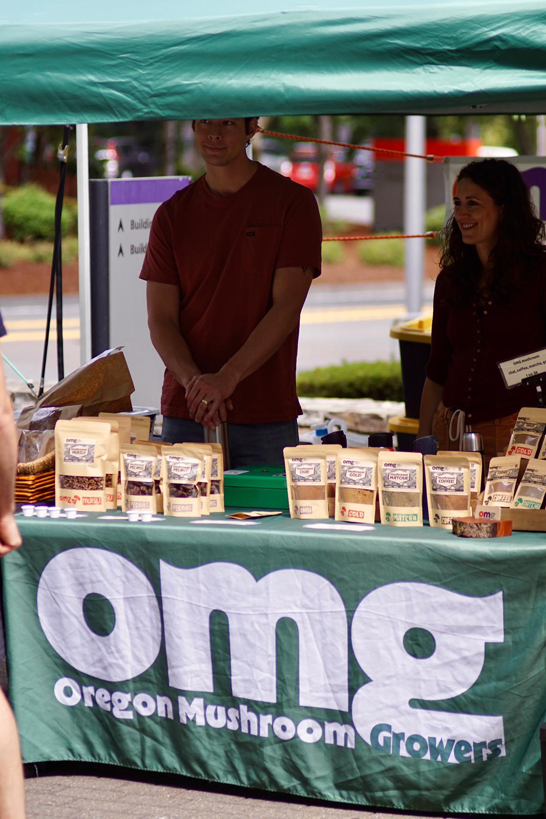 People behind a table with mushroom products at an outdoor market stall, green canopy overhead, Oregon Mushroom Growers banner visible.