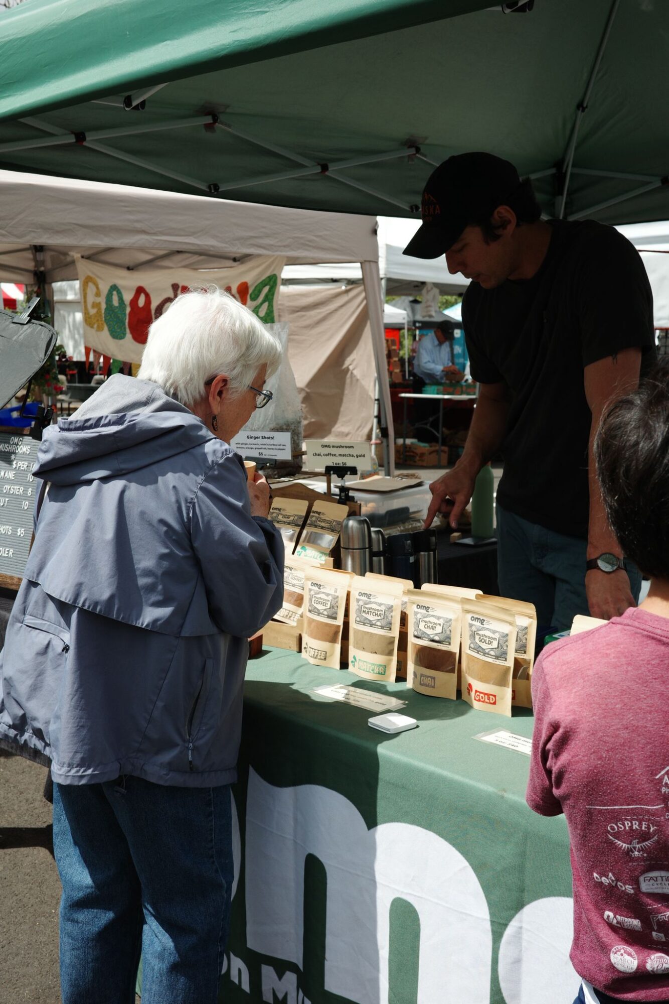Person with white hair and gray jacket talking to a vendor at an outdoor market stall, with various products displayed on the table.