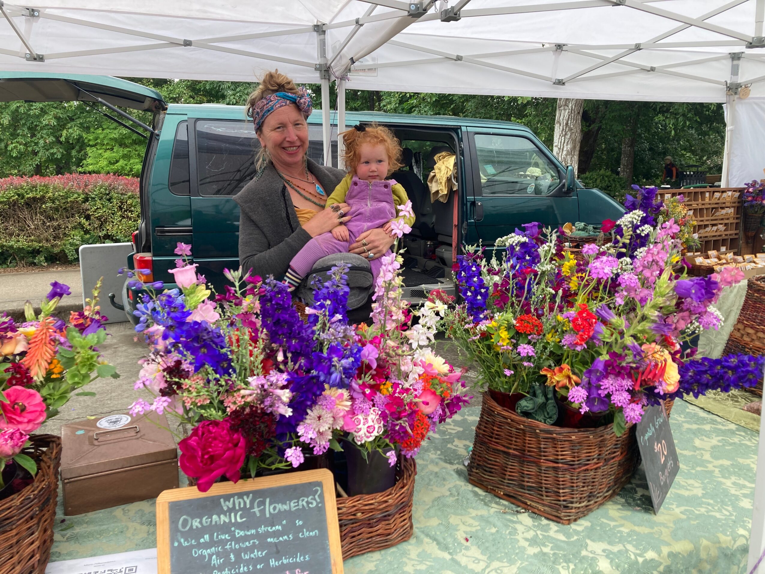 Woman holding a child at a flower stand with colorful flowers, a van, and a white canopy in the background.