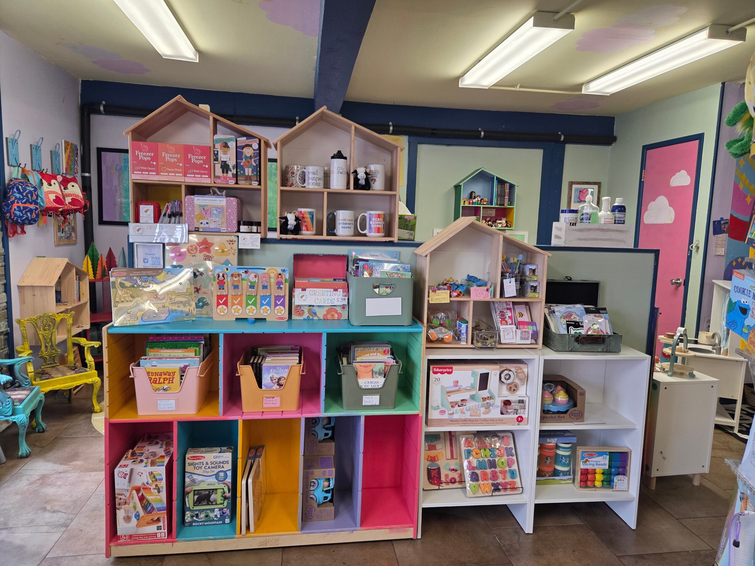 Colorful children's play area with shelves filled with toys, books, and games, in a bright room with ceiling lights.
