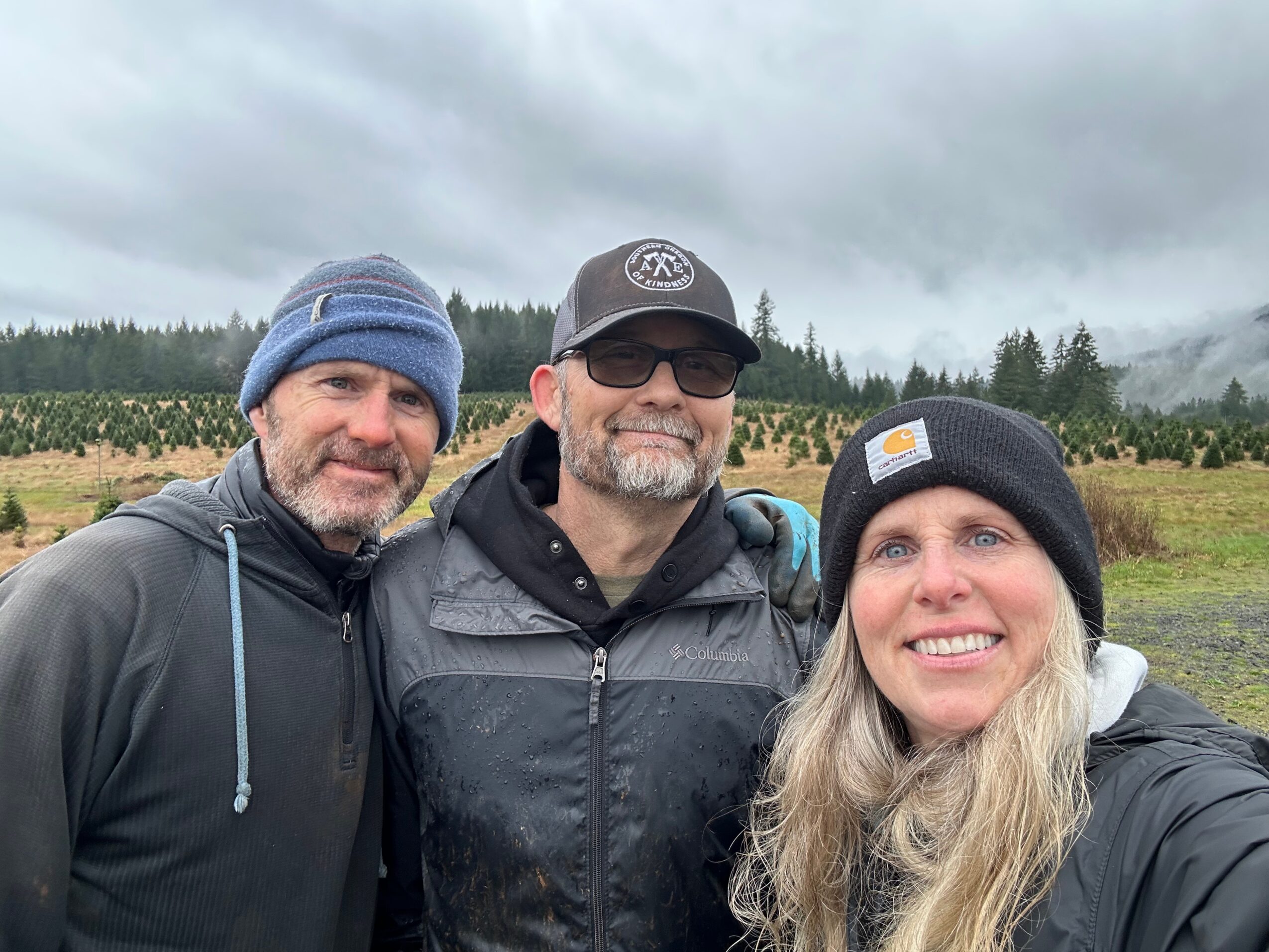 Three people outdoors on a cloudy day, smiling, with trees and fields in the background.