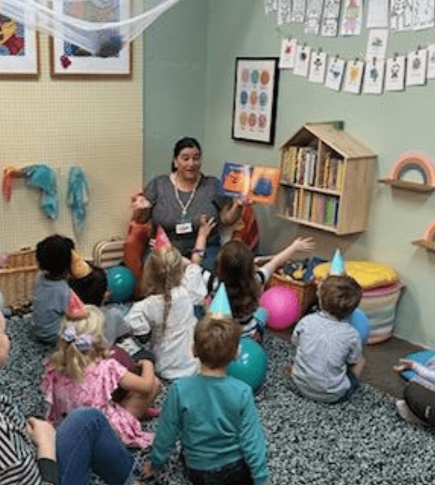 Teacher reading a book to children sitting on the floor in a classroom with colorful decorations and bookshelves.