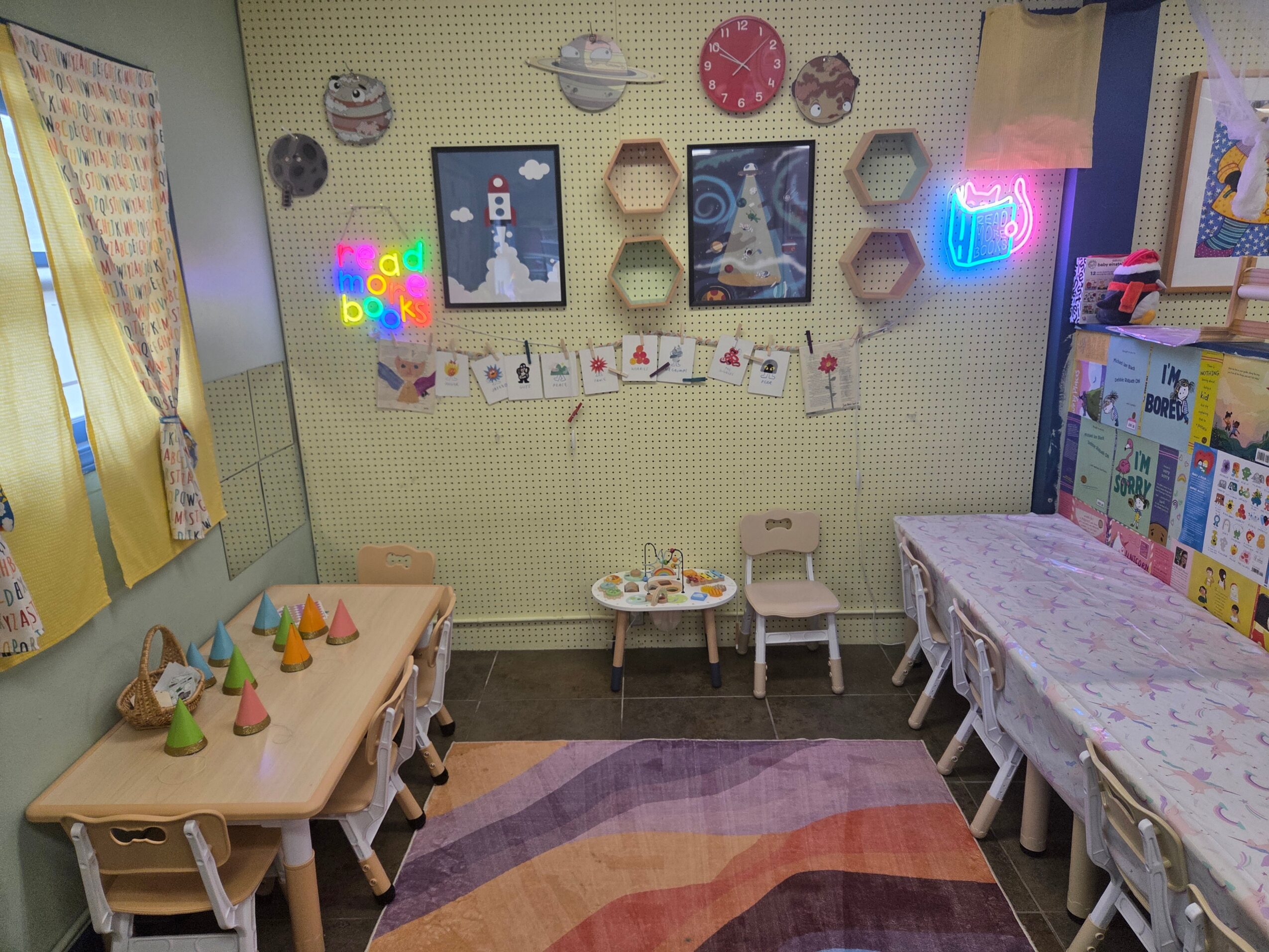 Classroom with tables, chairs, colorful decorations, and artwork on the wall, arranged for children, with a rug on the floor.