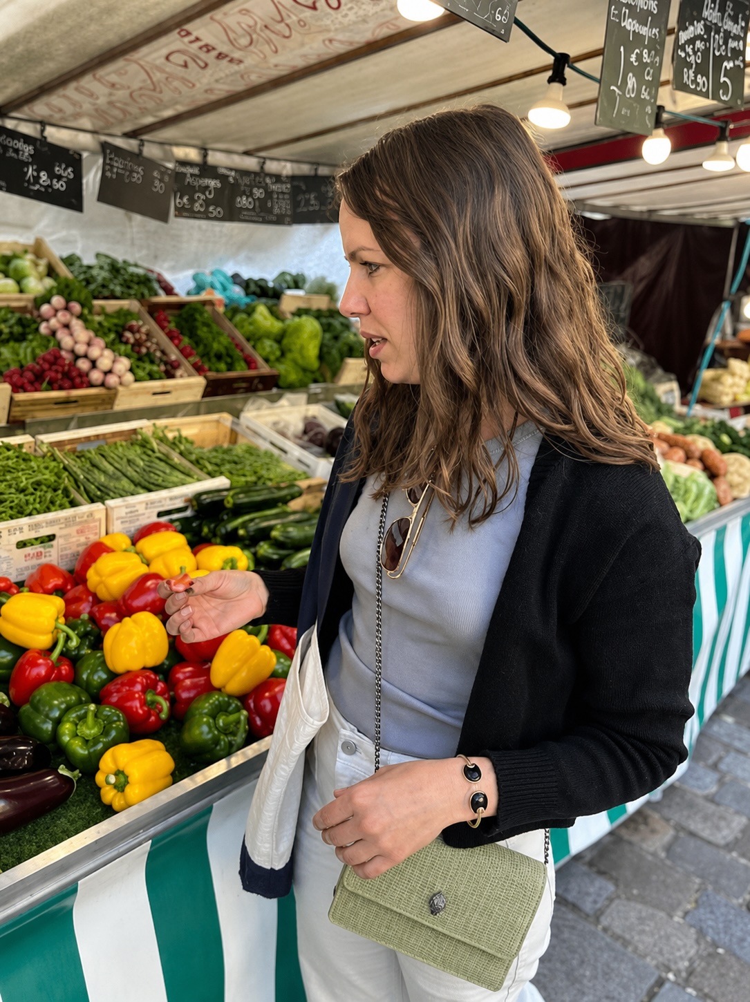 Woman shopping at a fruit stand with colorful vegetables and fruits, market stall with chalkboard signs overhead.