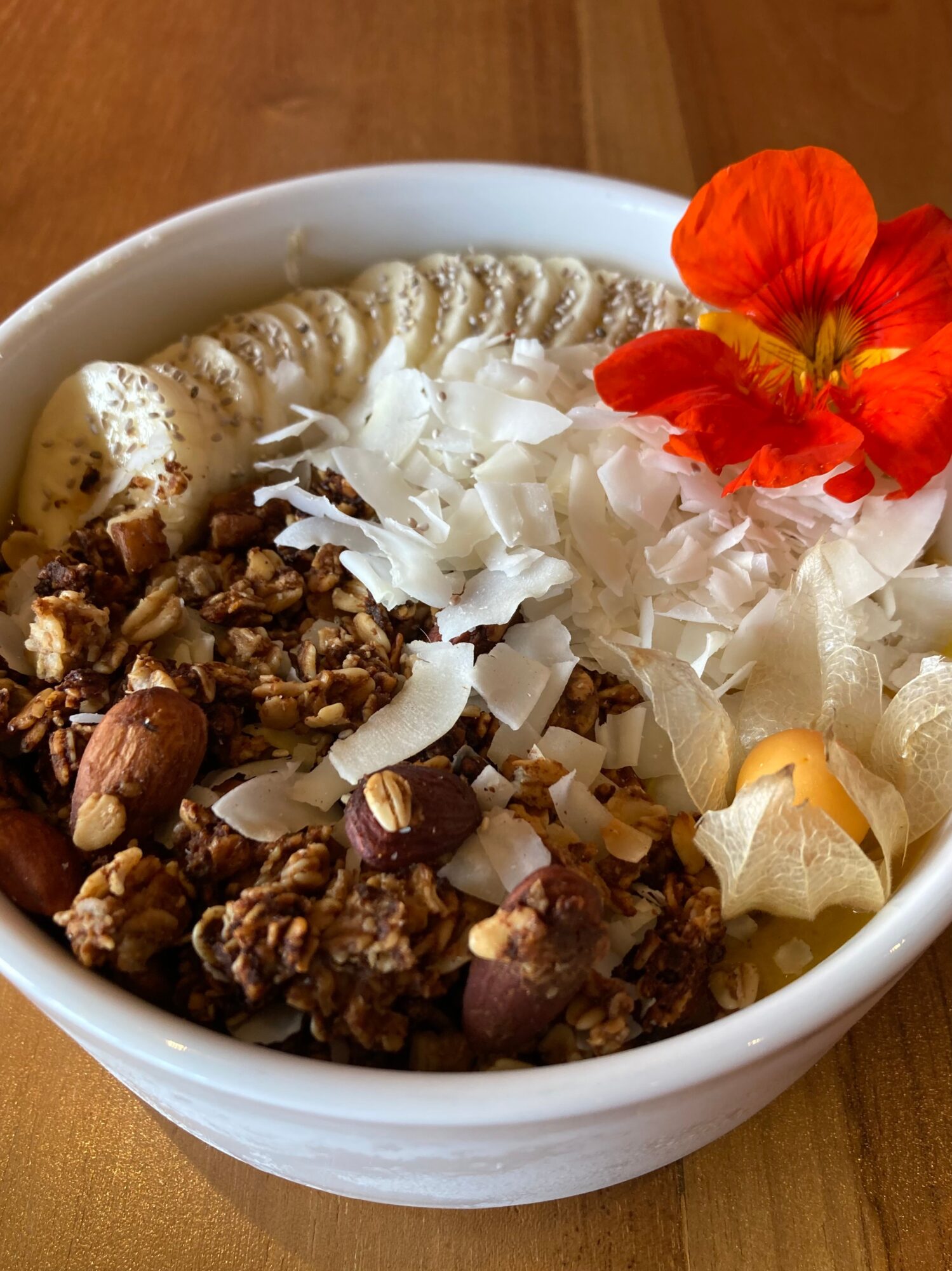 Bowl with yogurt, granola, coconut flakes, nuts, and an orange flower on top, on a wooden surface.