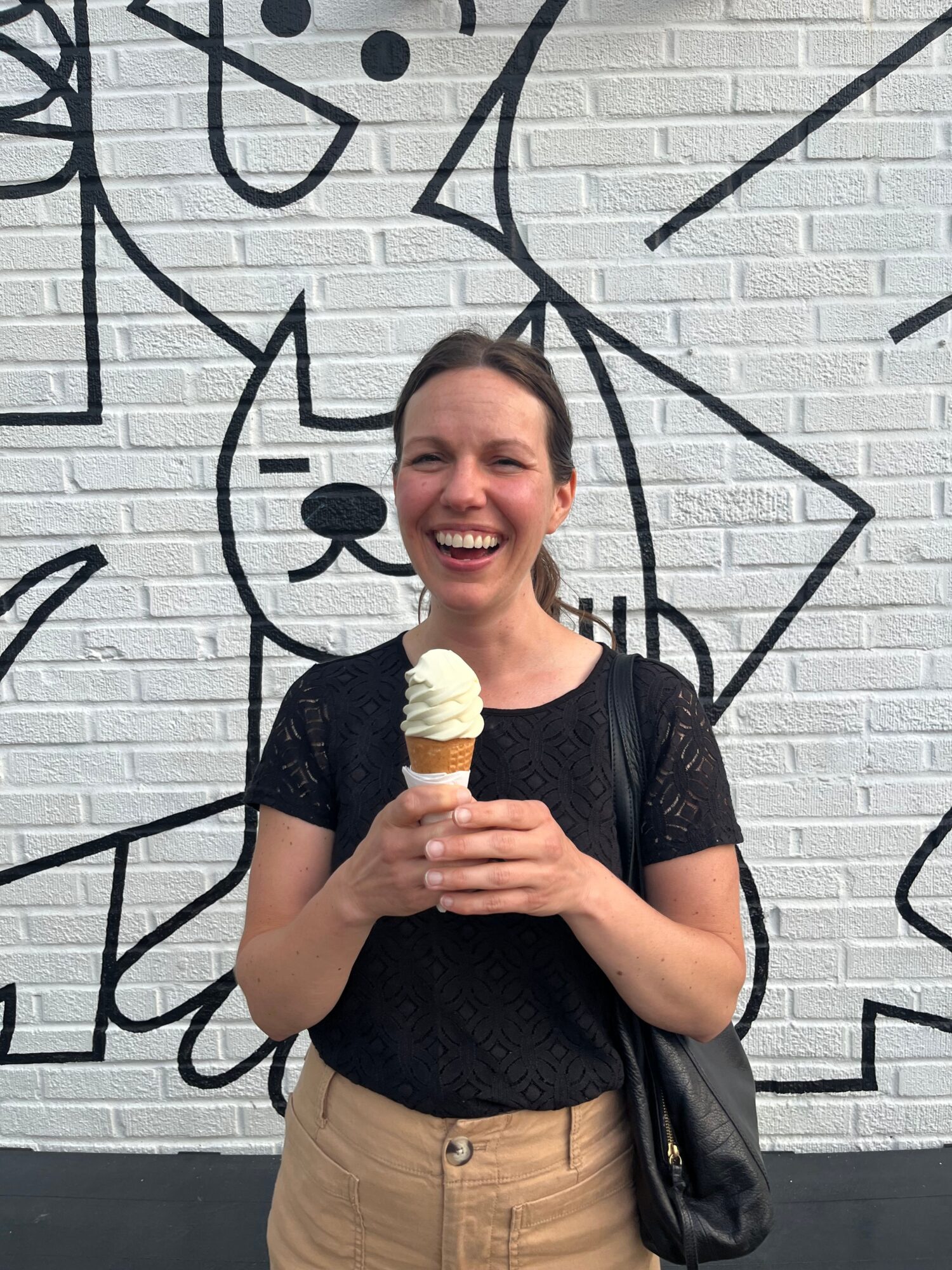 Woman smiling holding an ice cream cone, standing against a white brick wall with black line art, wearing a black top and beige pants.