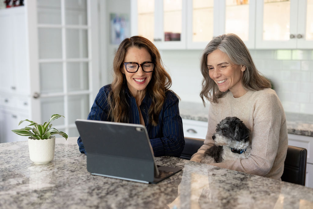 Two women smiling at a laptop in a bright kitchen, one holding a small dog, with a potted plant nearby.