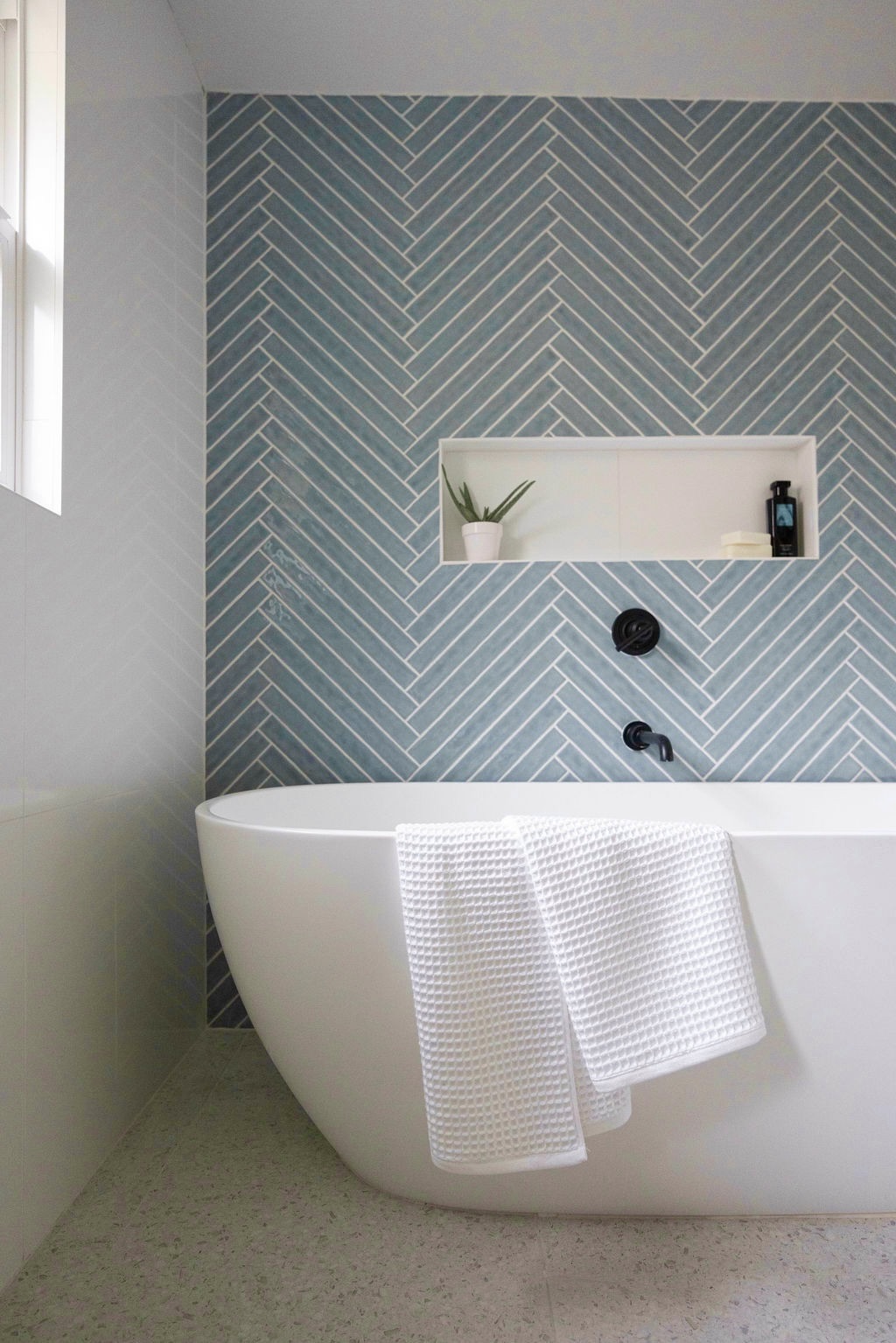 Bathtub with a white towel hanging over the edge, blue herringbone tile wall, small shelf with plant and bottle, window on left