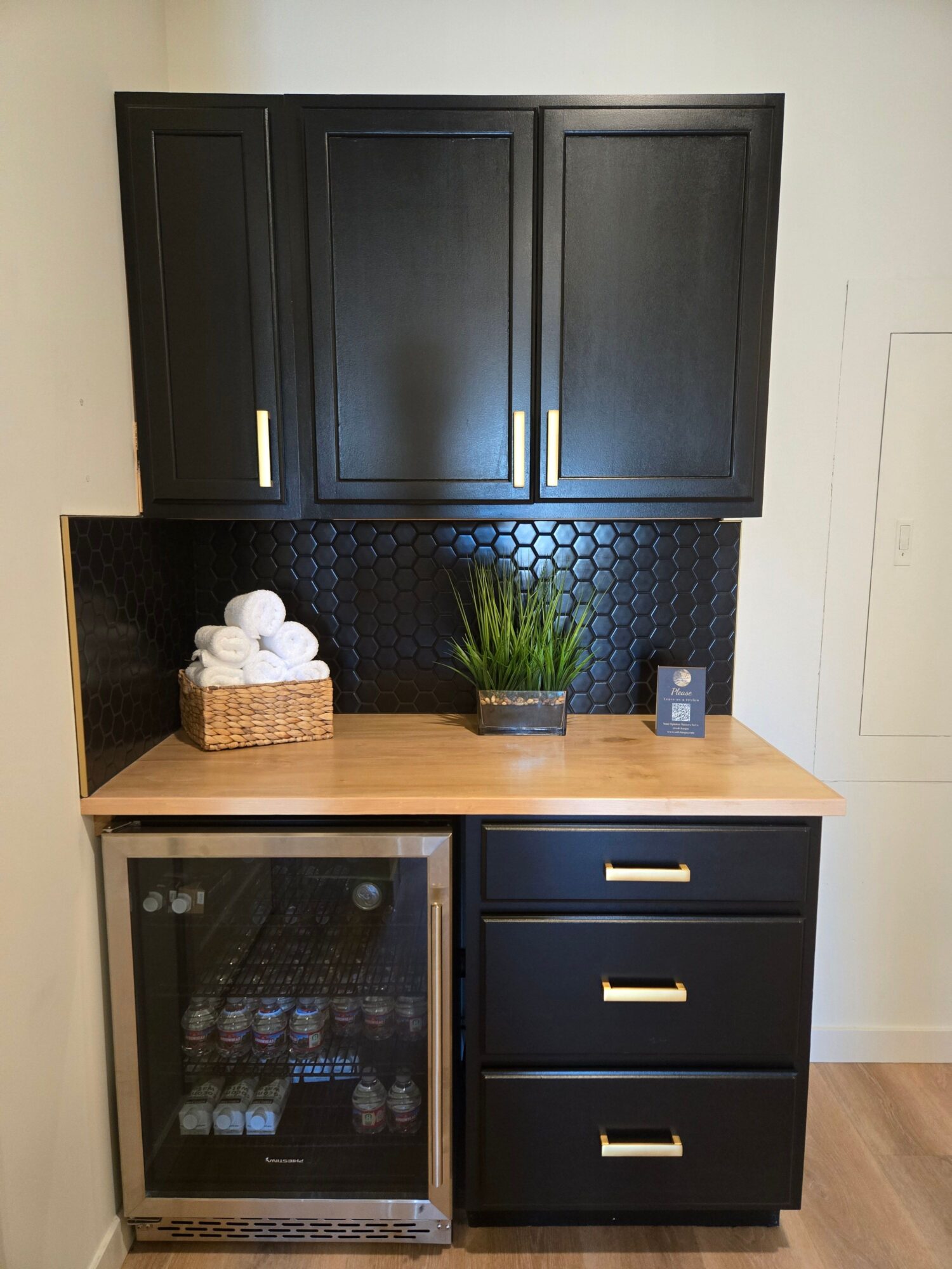 Black cabinets, a small plant, and a mini fridge in a kitchen corner, with a wooden countertop and black backsplash.