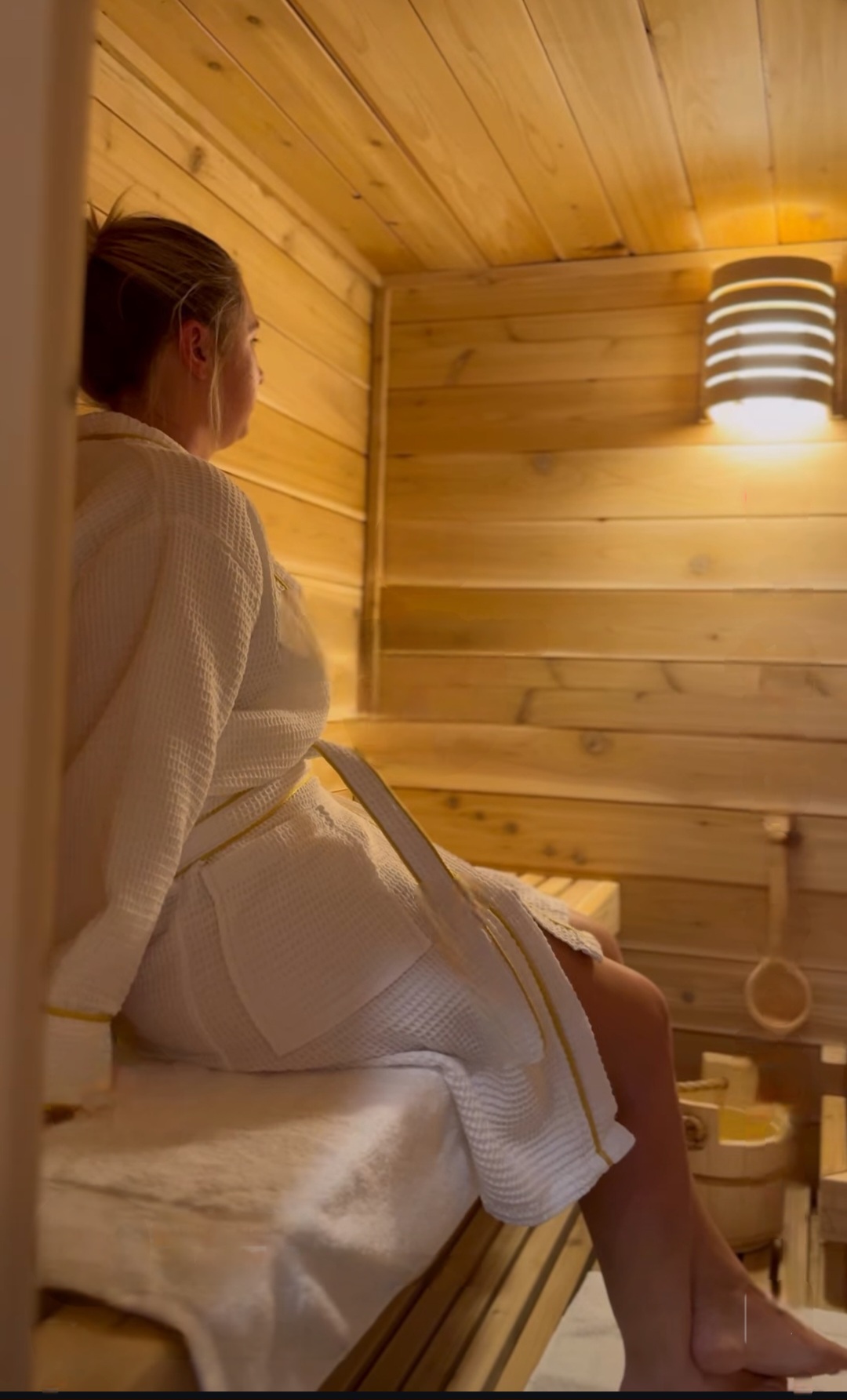 Person sitting on a wooden bench in a sauna, wearing a white robe, with wooden walls and a light fixture.