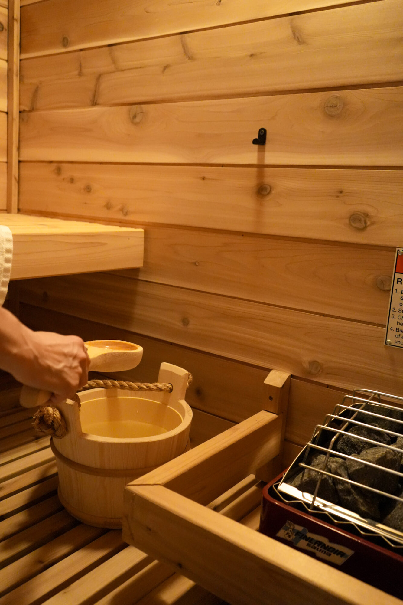 Wooden interior with a person holding a ladle near a wooden bucket and a grill, warm lighting, wooden walls and floor.