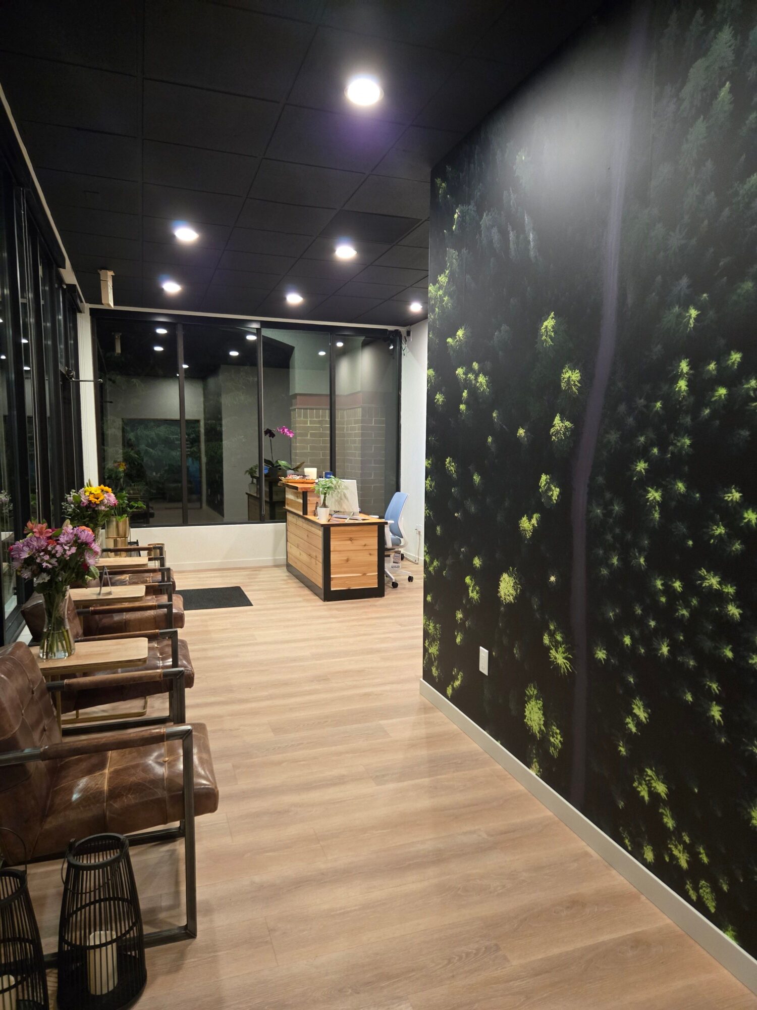 Room with dark ceiling, wooden floor, large window, and a wall with a nature scene, furniture, and flowers.