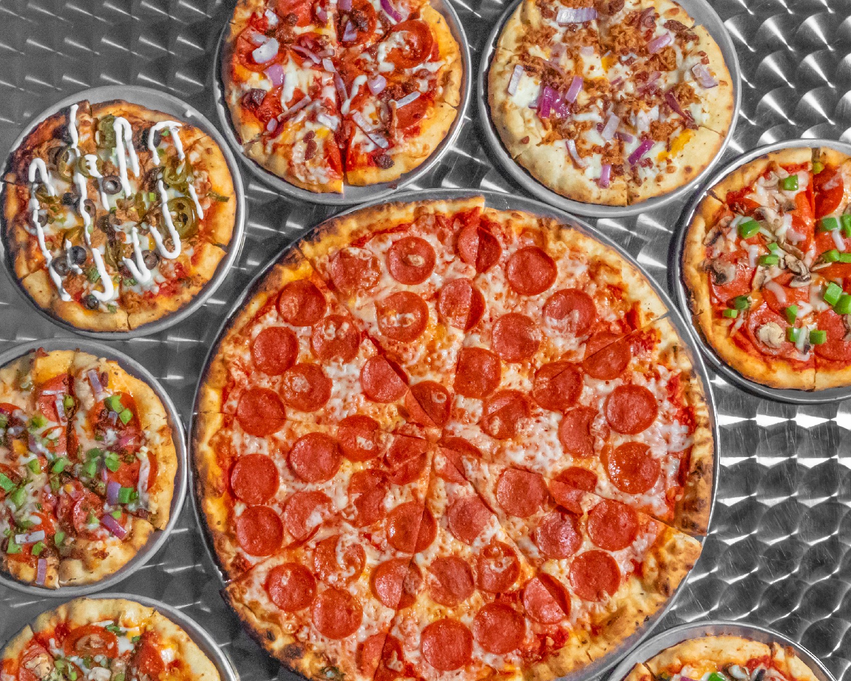 Multiple pizzas on a metal surface, with one large pepperoni pizza in the center and smaller pizzas around it.