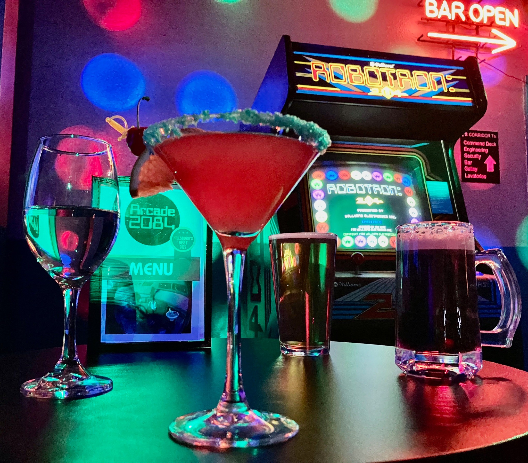 Bar with colorful drinks, arcade game, and neon signs, illuminated by vibrant lights.