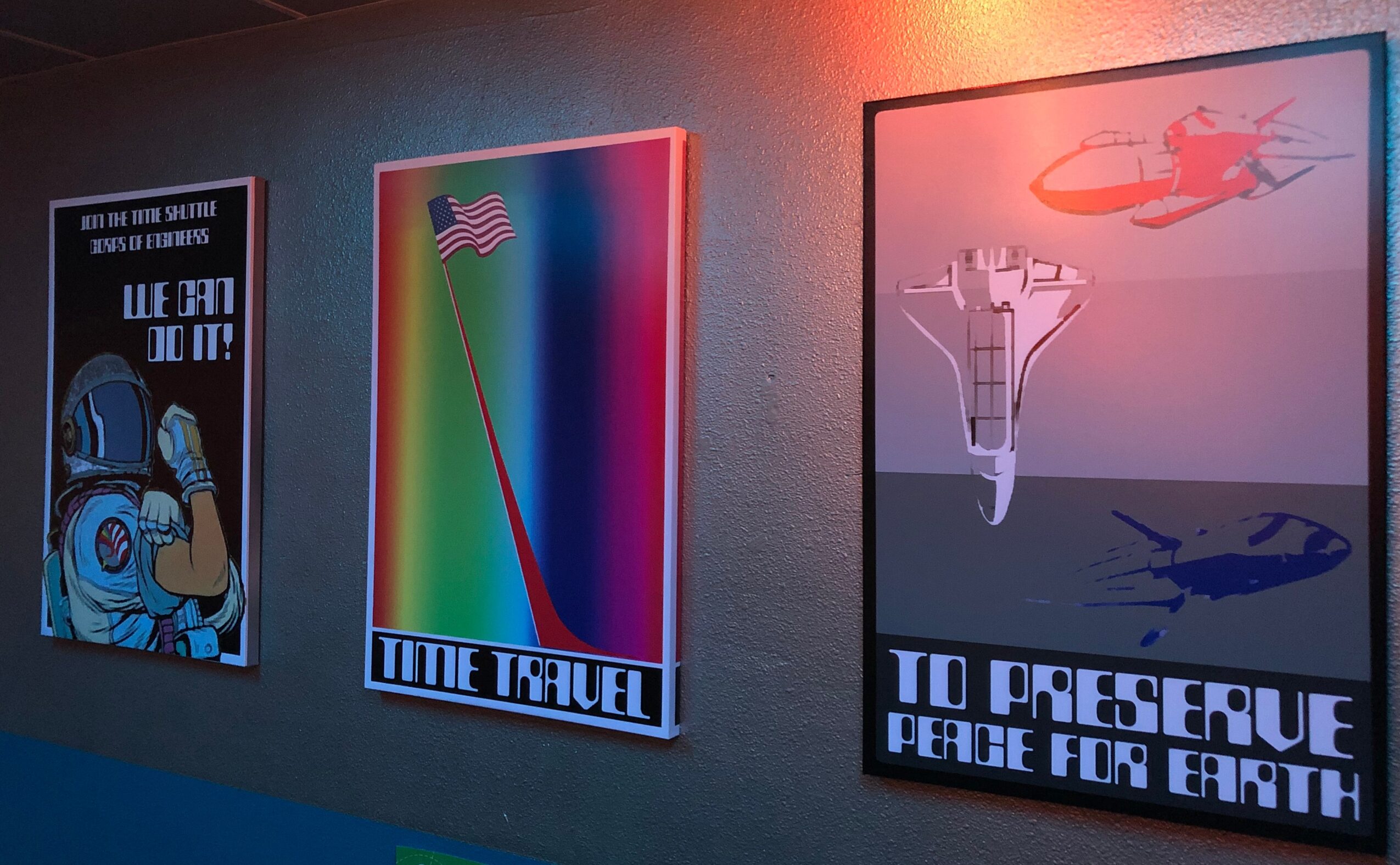 Three posters on a wall, one with astronaut and text, one with rainbow and flag, one with helicopter and airplane, all with text.