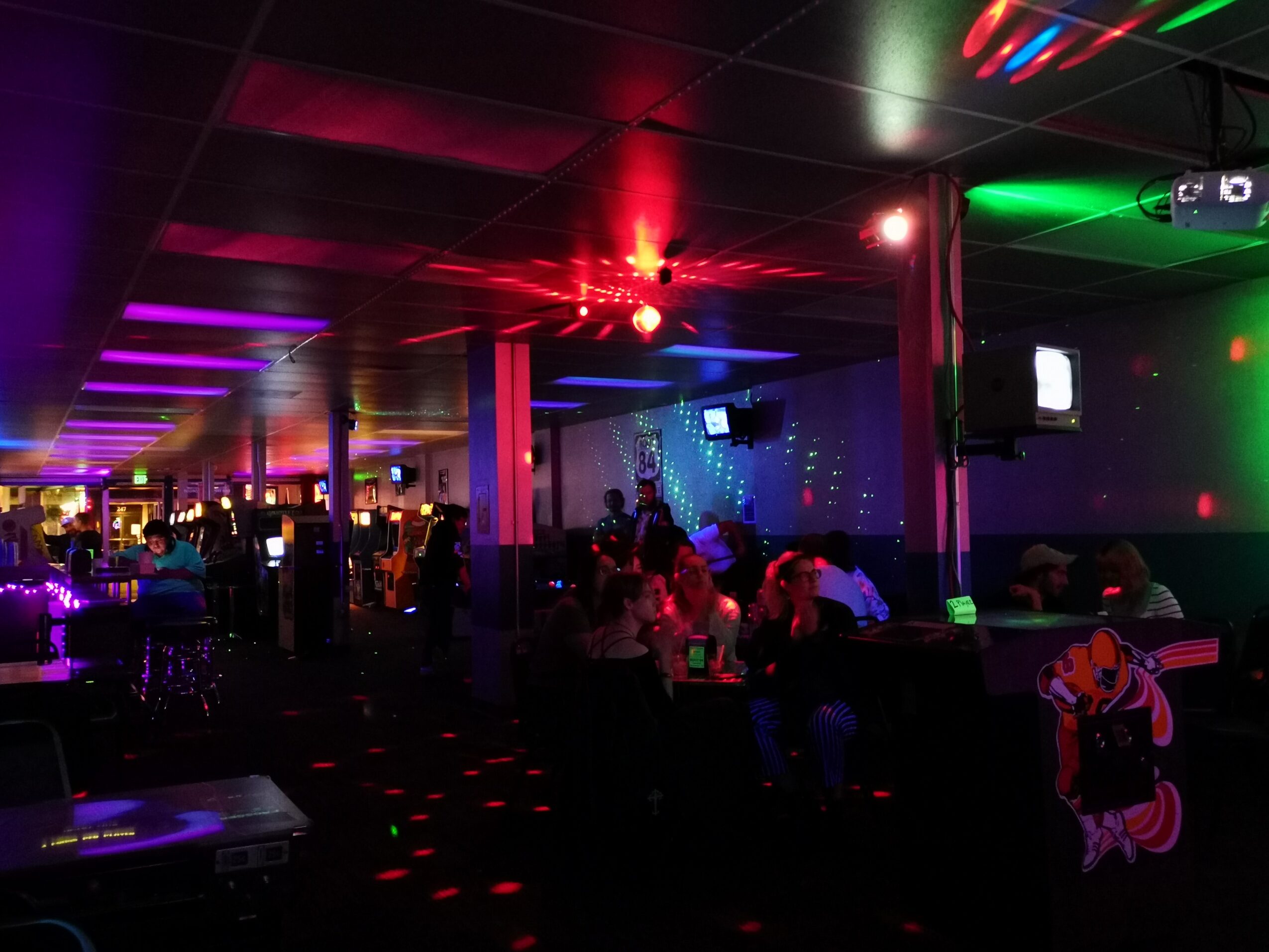 Nightclub interior with colorful lights, people sitting and socializing, and a dance floor with illuminated patterns.