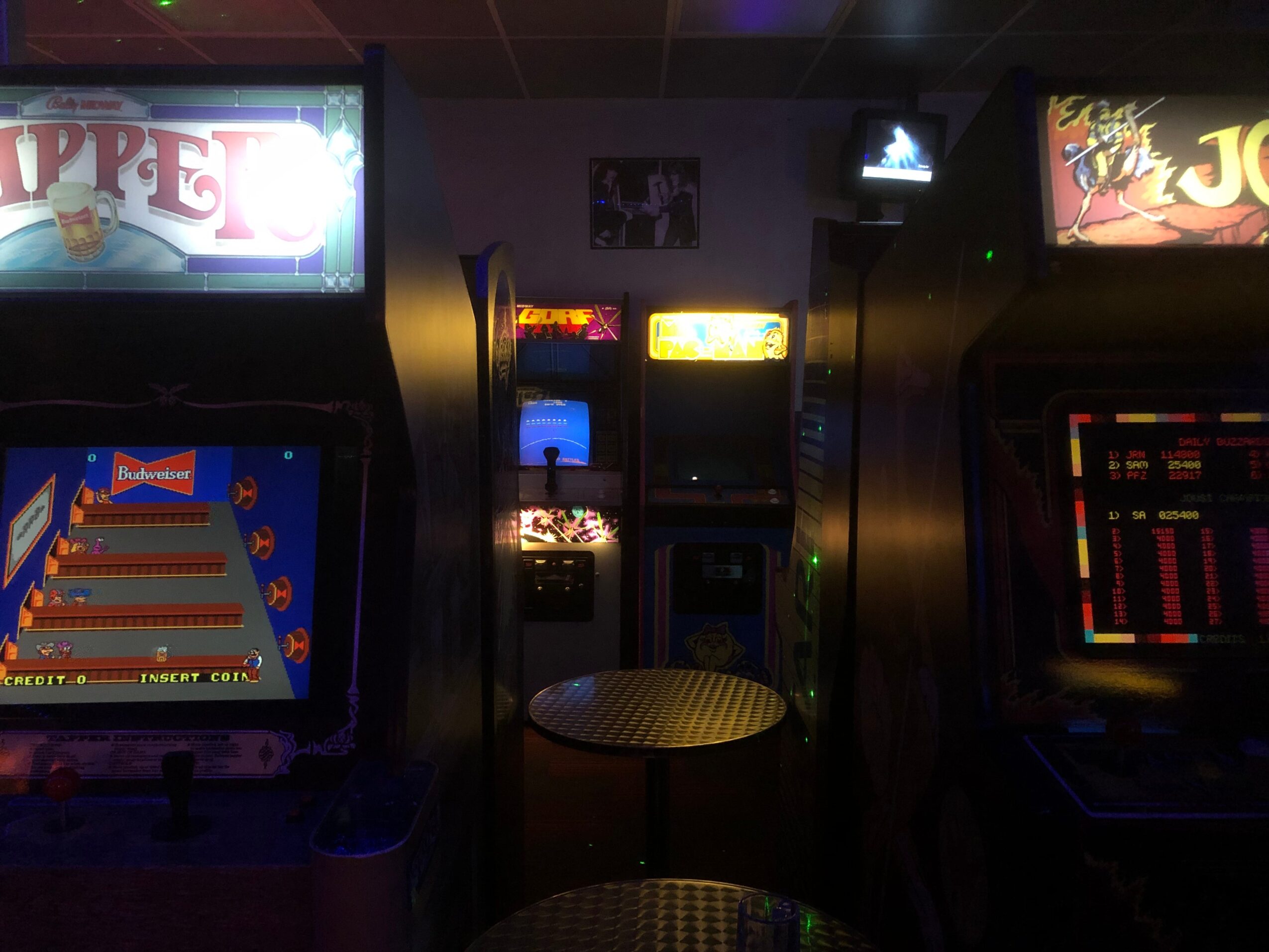 Dark room with illuminated arcade game screens, a small round table, and a framed picture on the wall.