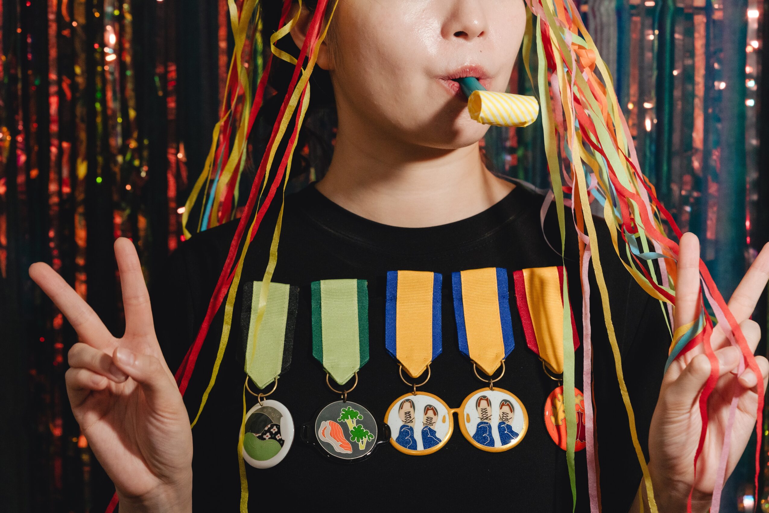 Person with colorful streamers in hair, wearing medals, holding a party horn, making peace signs with both hands.