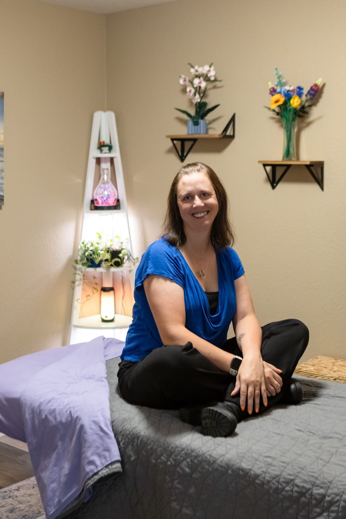 Woman sitting cross-legged on a bed in a room with wall shelves and flower arrangements, smiling at the camera.