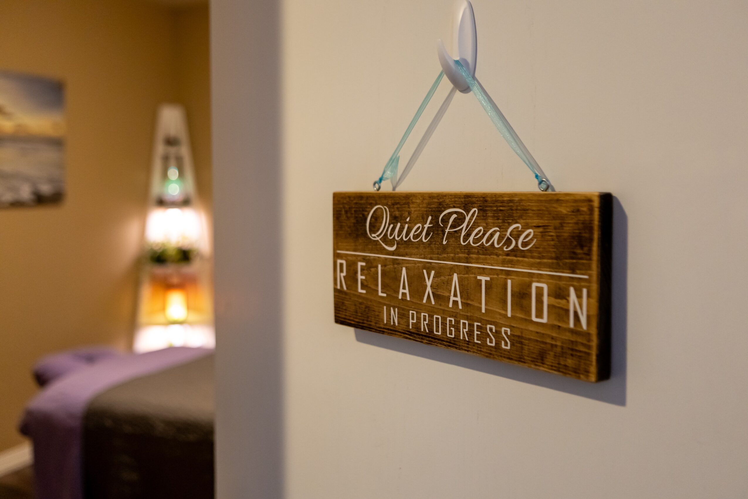 Wooden sign hanging on a wall reads 'Quiet Please Relaxation in Progress' with a blurred bedroom in background.