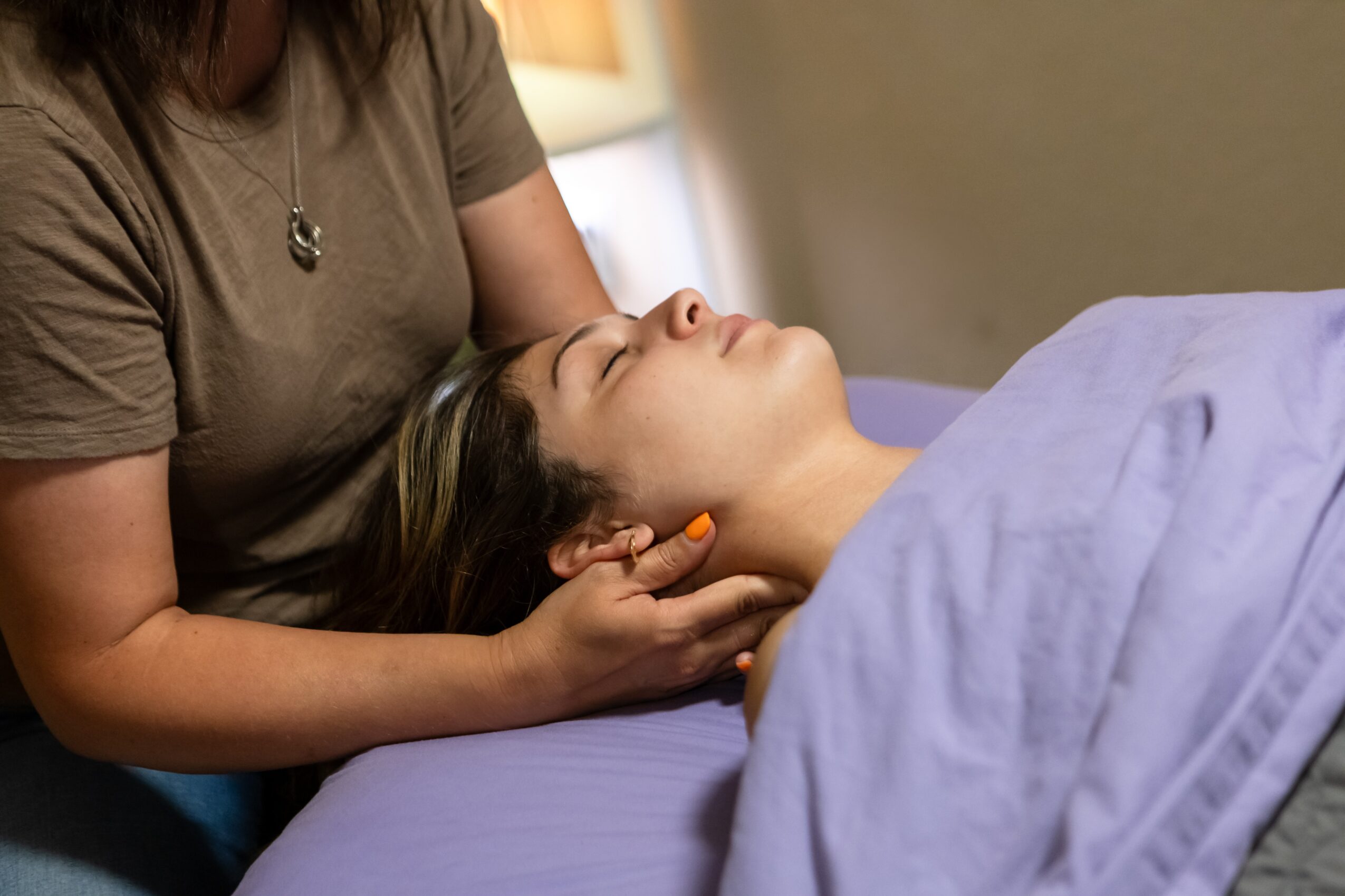 Person receiving a neck massage while lying on a bed, with a caregiver supporting their head, in a room with warm lighting.