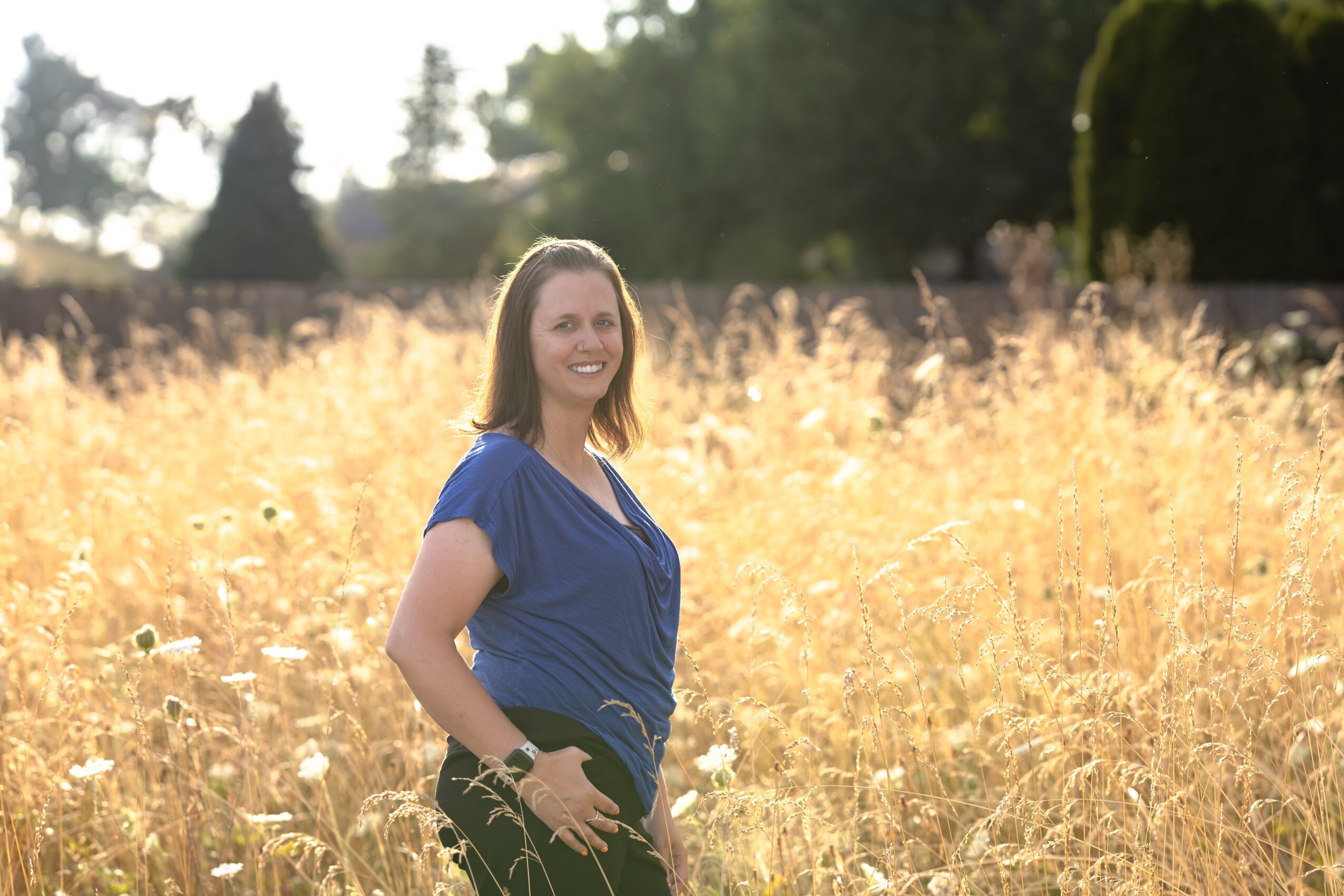 Woman standing in a field of tall, golden grass, smiling, with trees and sky in the background.
