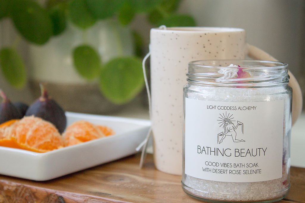 A lit candle in a glass jar with a label, a white mug, and a plate of oranges on a table, with green plants in the background.