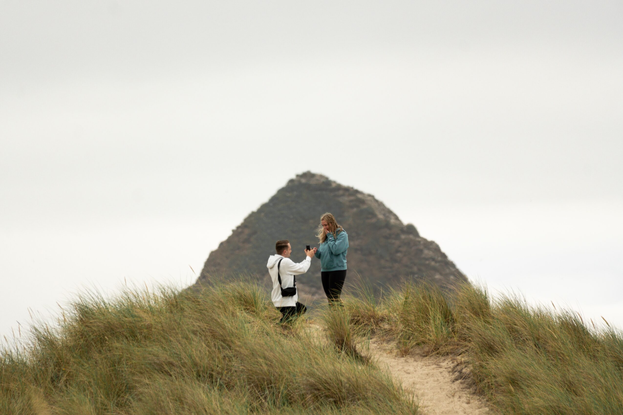Two people stand on grassy terrain with a mountain in the background, engaging in conversation or an activity outdoors.