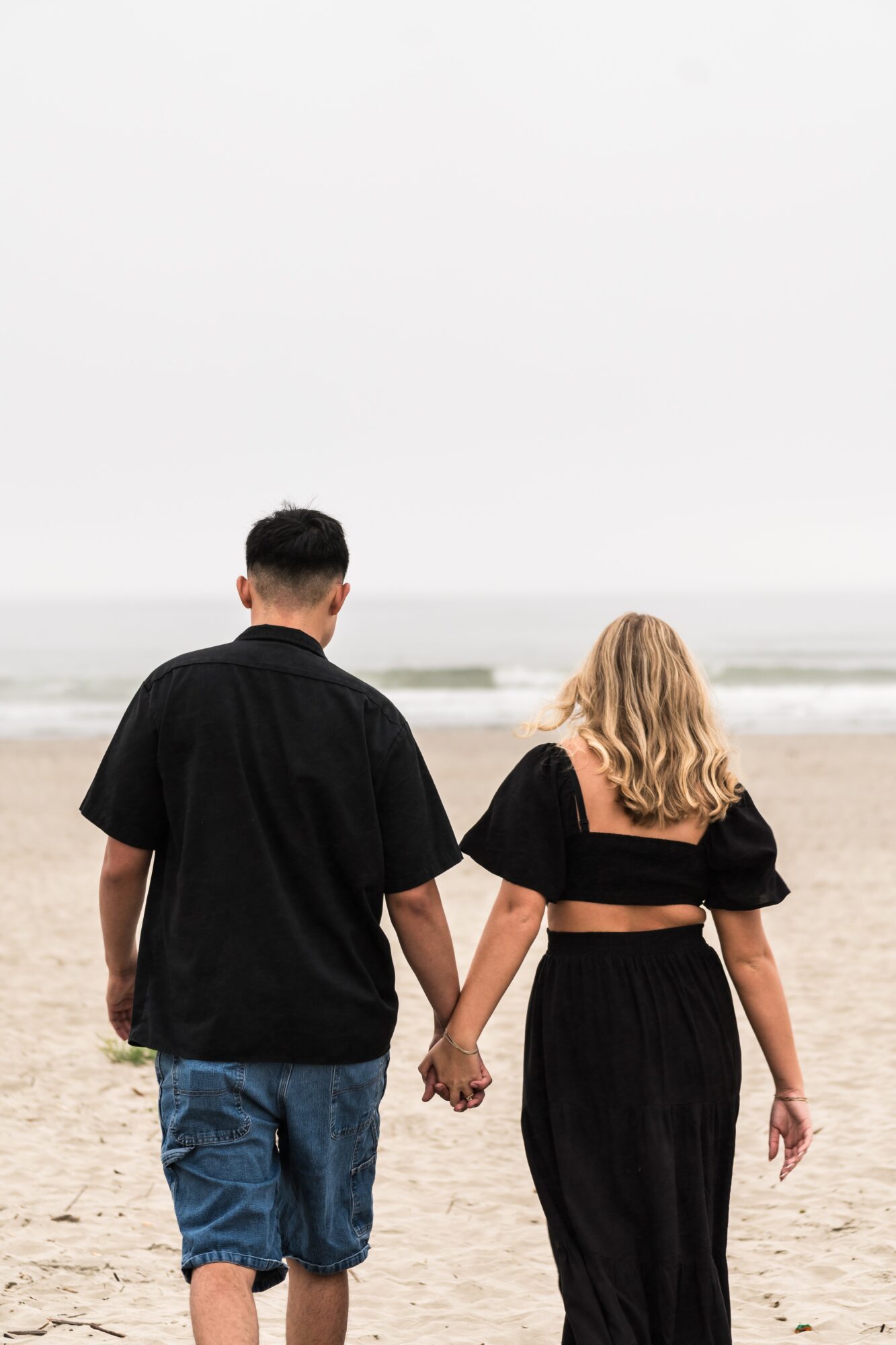 Two people holding hands walk on the beach, facing the ocean, with their backs visible, overcast sky, sandy shore.