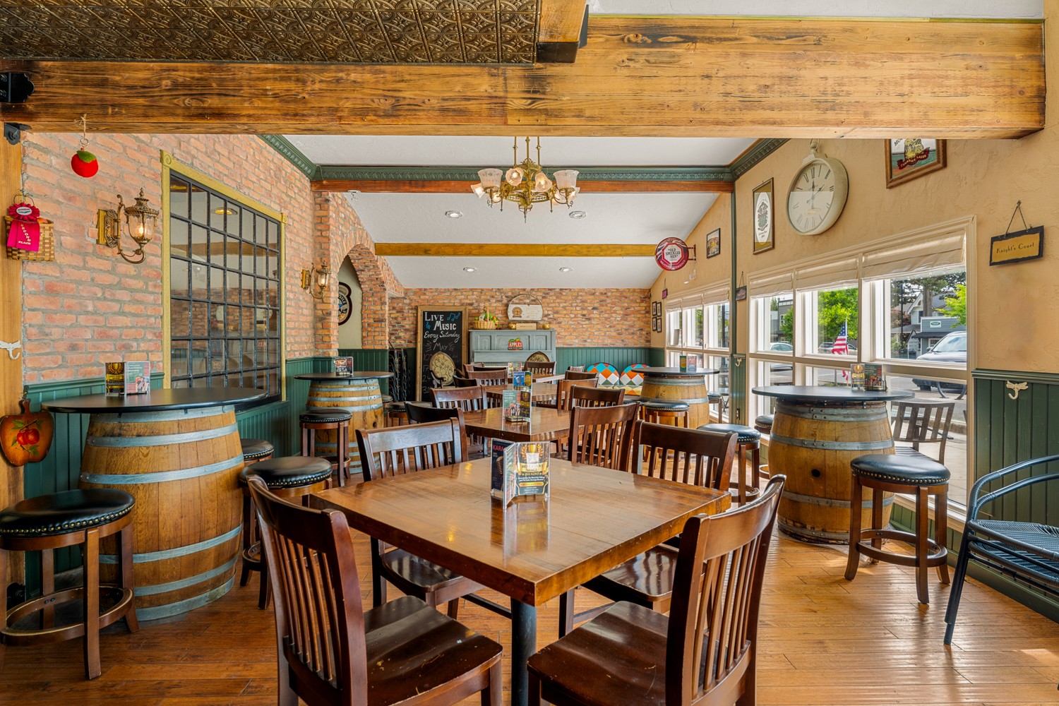 Interior of a restaurant with wooden tables, chairs, barrels, large windows, and a chandelier, bright and inviting atmosphere.