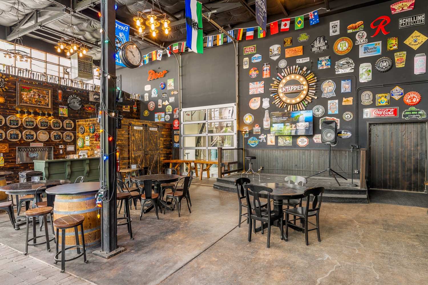 Interior of a bar or cafe with tables, chairs, and a decorated wall with signs and flags, illuminated by hanging lights.