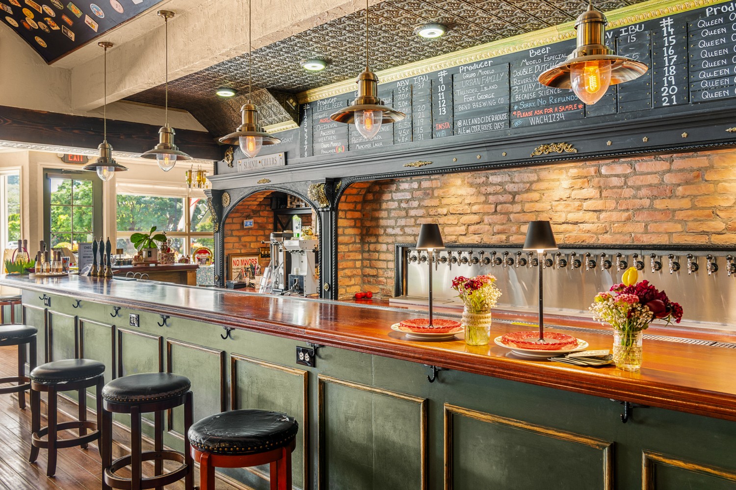 Interior of a cafe with a wooden counter, bar stools, brick wall, menu boards, and hanging lights, with windows showing outdoor scenery.