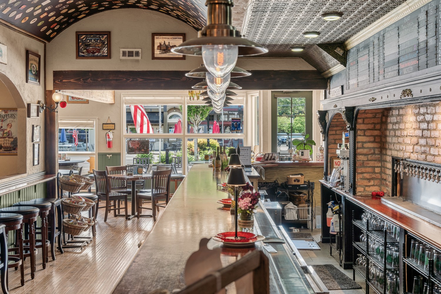 Interior of a restaurant with a long wooden bar, tables, chairs, and large windows, decorated with artwork and hanging lights.