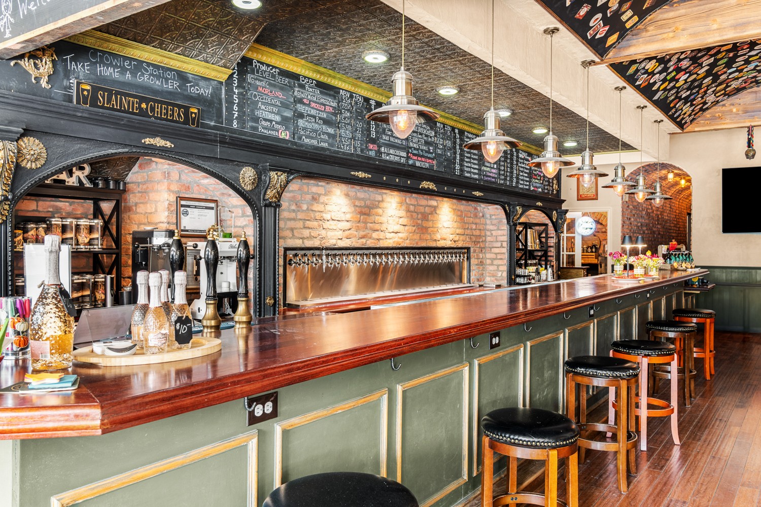 Bar with a long wooden counter, bar stools, and a chalkboard menu above. Brick walls and decorative ceiling elements visible.