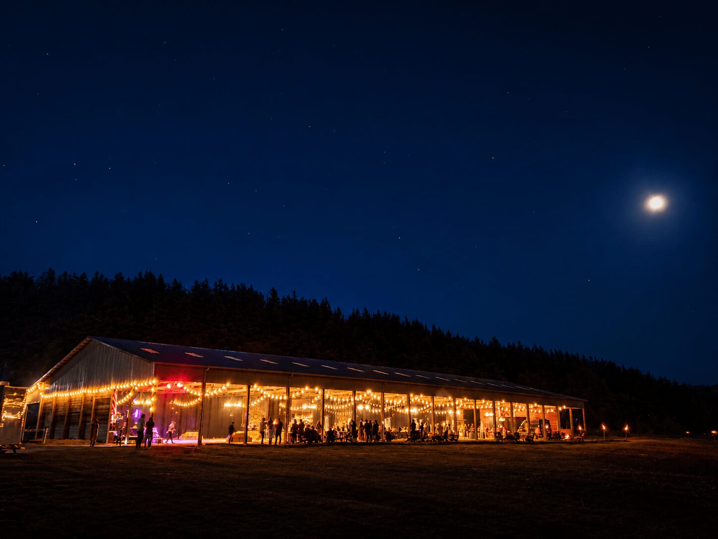 Night scene with a large, illuminated tent and people inside, under a starry sky with a bright moon.
