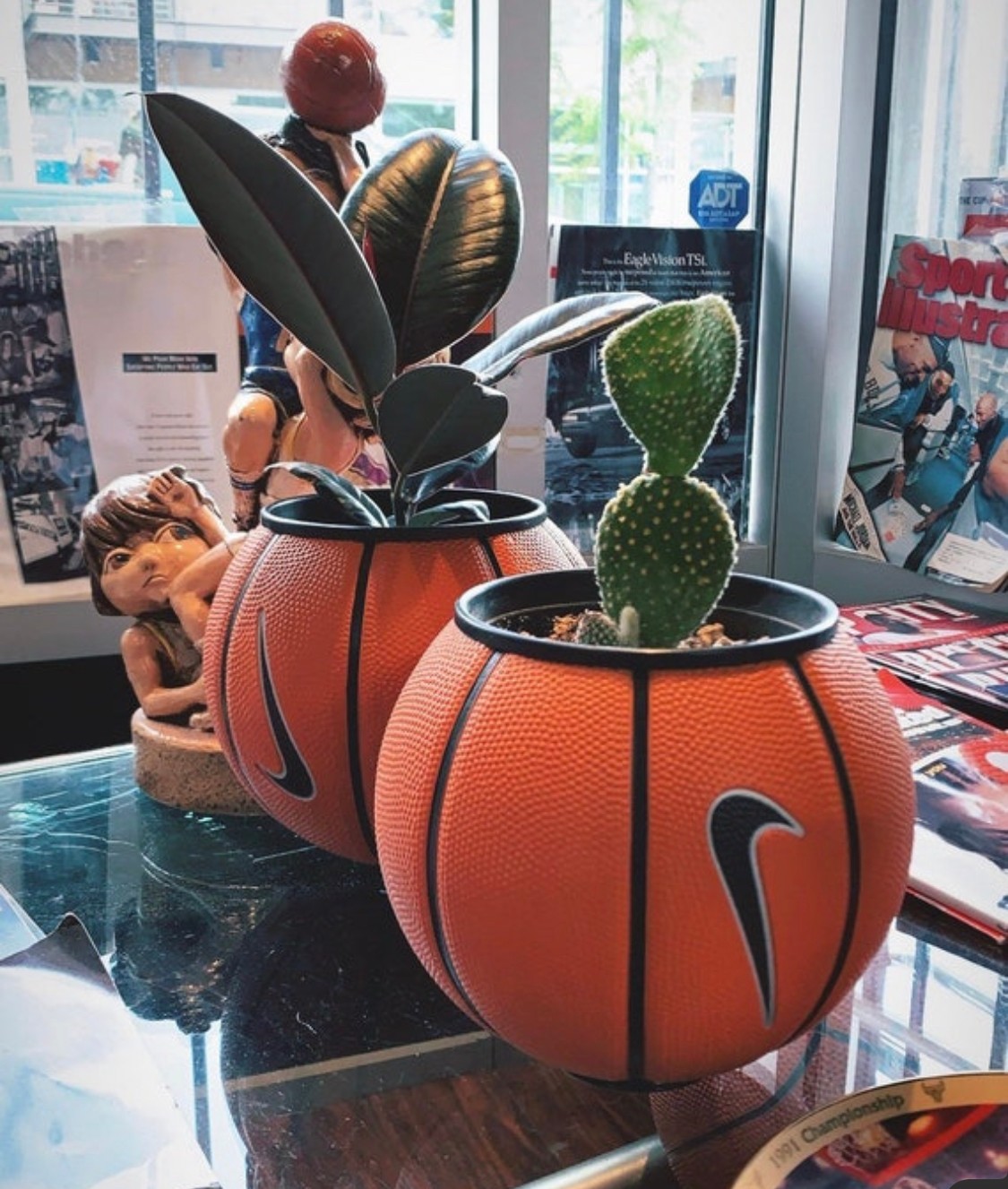 Two orange basketball-shaped planters with green plants inside, placed on a reflective surface near a window, with magazines and a doll in the background.