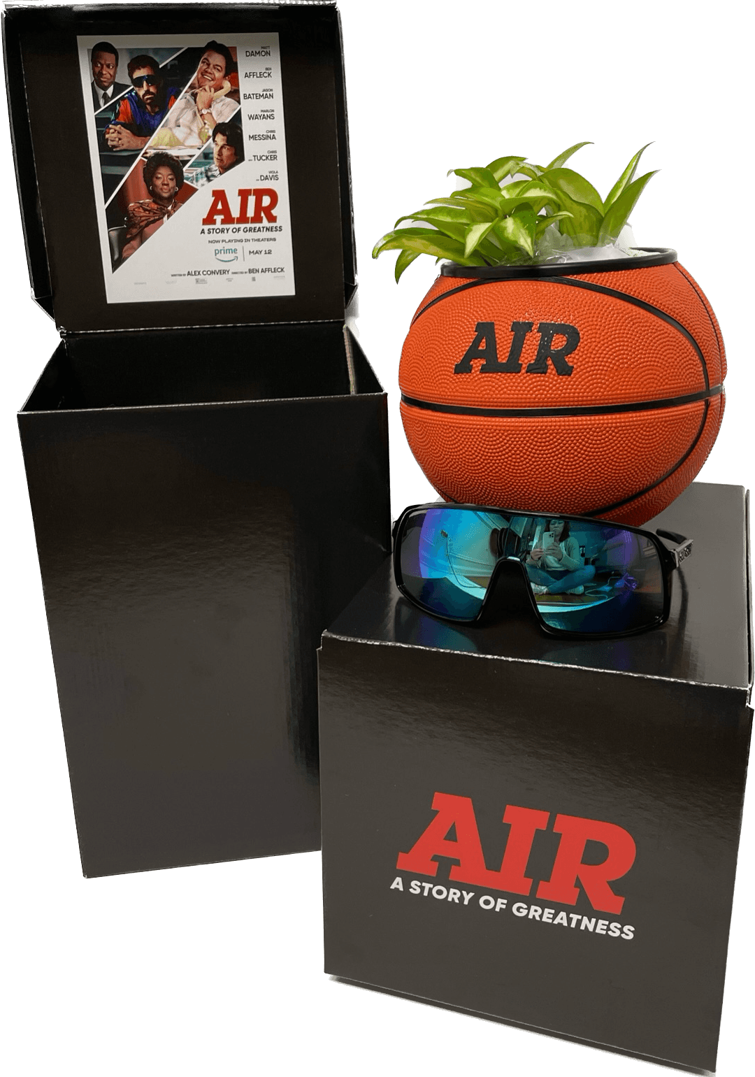 A basketball-shaped planter with green plants, sunglasses, and a movie poster in a black box, with text 'AIR' and 'A STORY OF GREATNESS'.