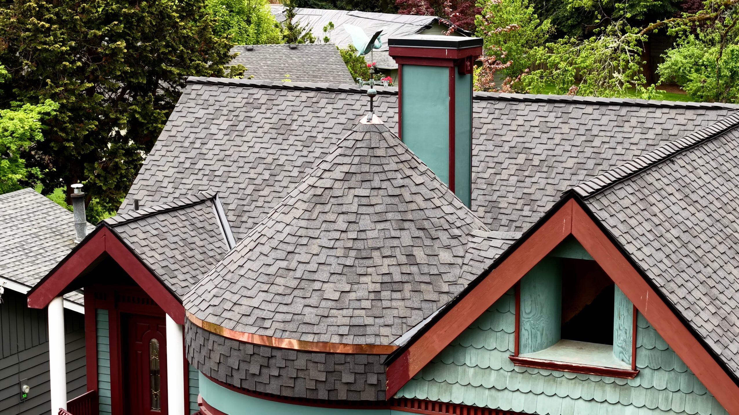 Roof with multiple gables, a chimney, and green siding, surrounded by trees, viewed from above.