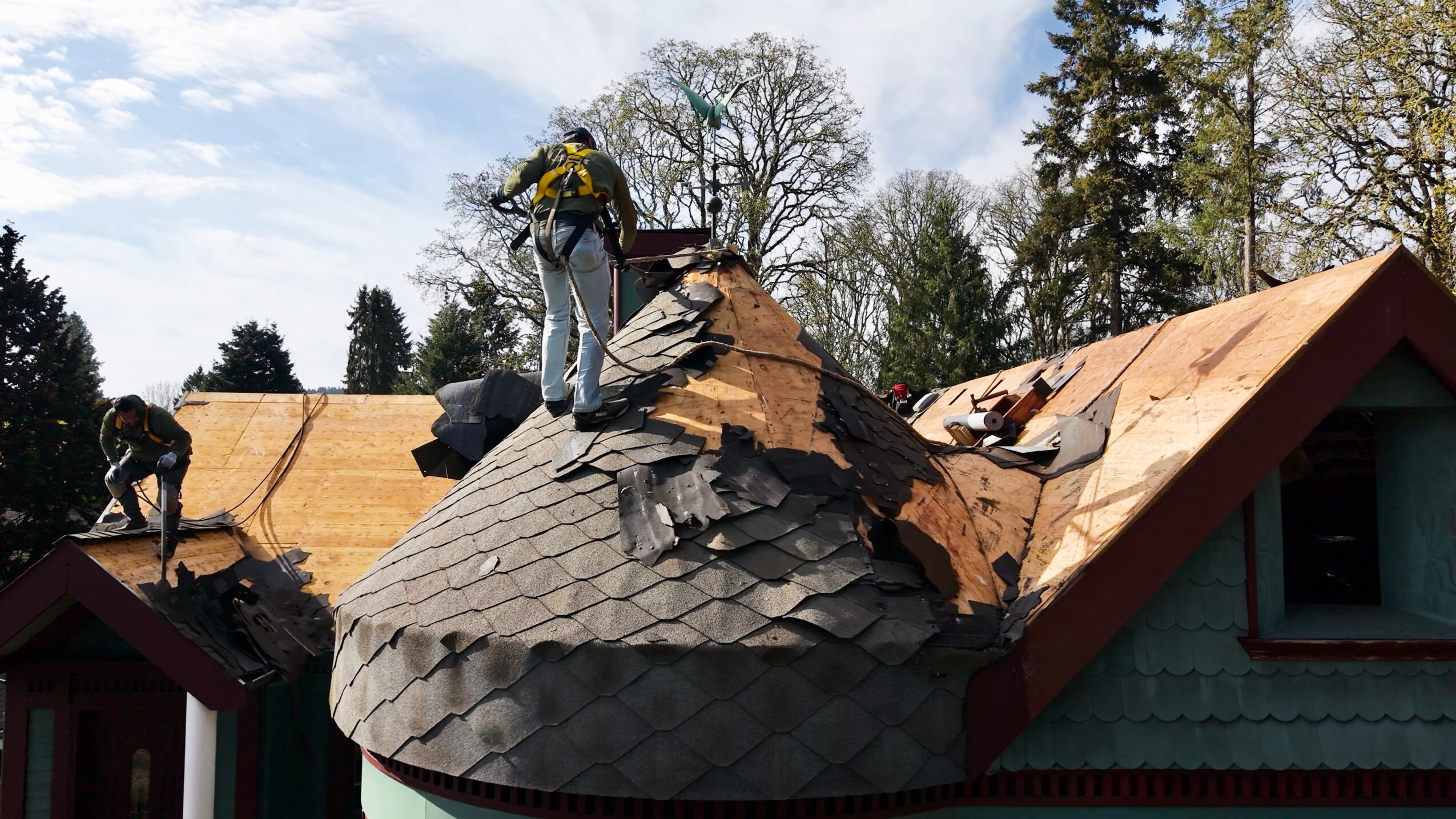 Workers repairing a damaged roof with broken shingles and a chimney, trees and a blue sky in the background.