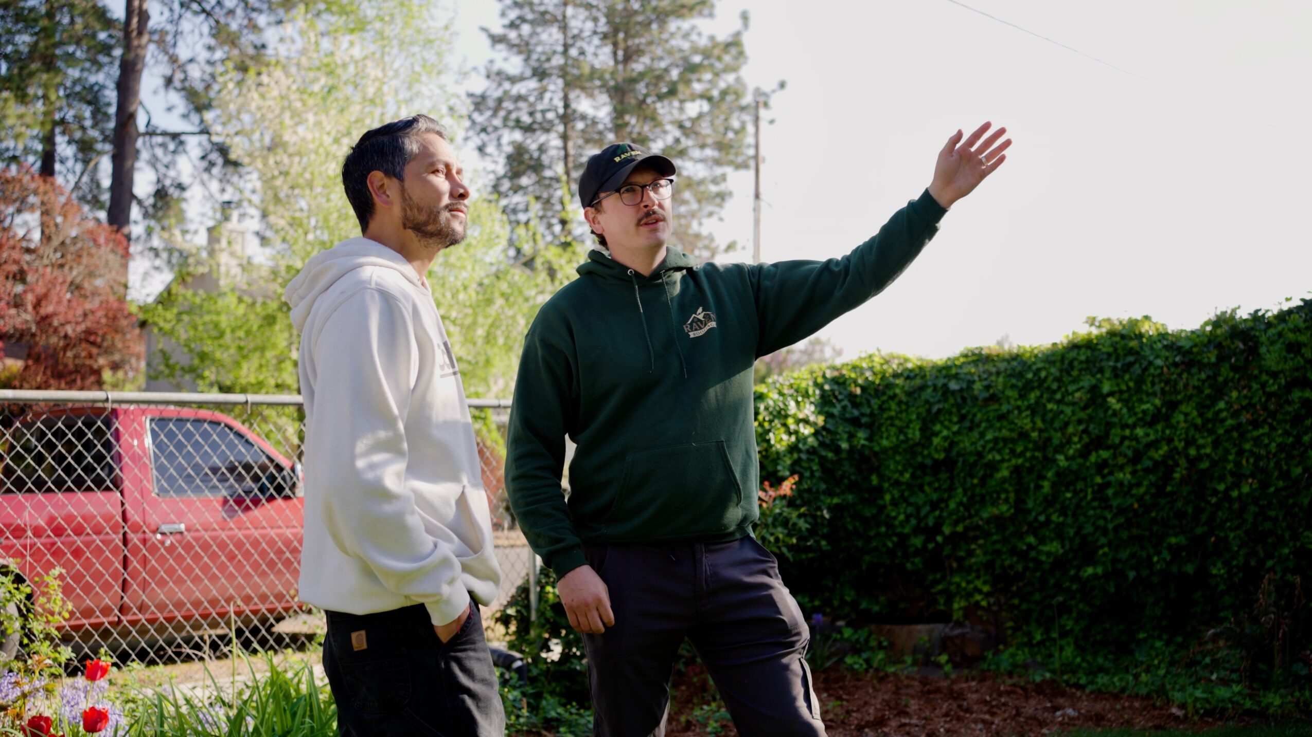 Two men standing outdoors near a hedge, one gesturing with his hand, trees and a red vehicle in background.